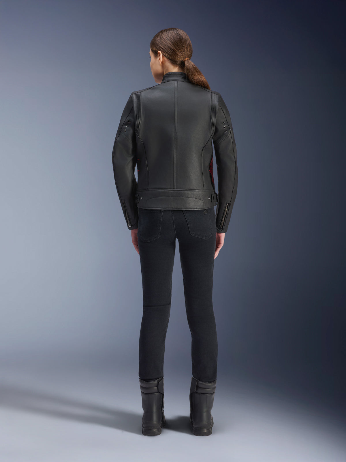 Ryggvy av en kvinna som bär Alpinestars Stella Dyno läderjacka, svart med Haute Red-detaljer, matchad med svarta slim-fit-byxor och svarta stövlar, modern heritage-design för motorcykeljacka i läder.