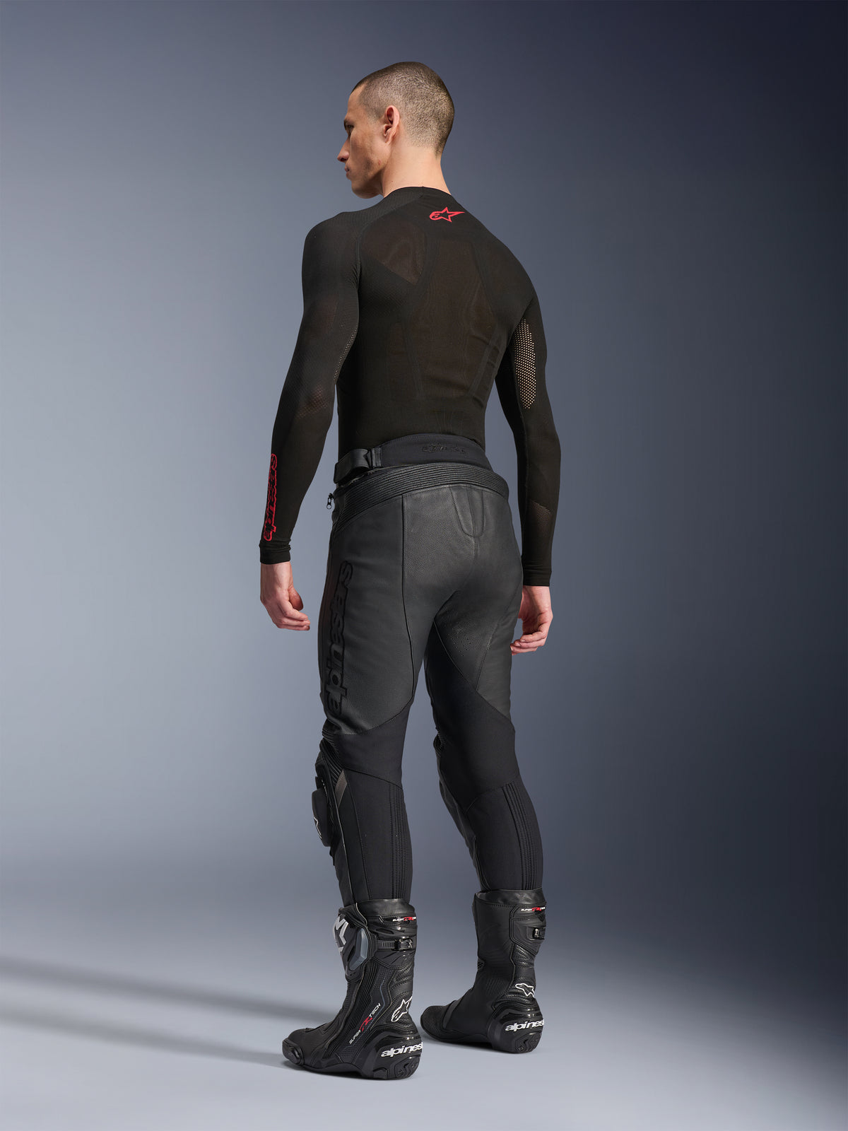 Alpinestars Missile V3 läderbyxor, svarta, sedda bakifrån på modell, bärs med ett svart långärmat underställ och svarta racingstövlar, med dragspelspaneler i läder och en förböjd design för en ergonomisk körpassform.