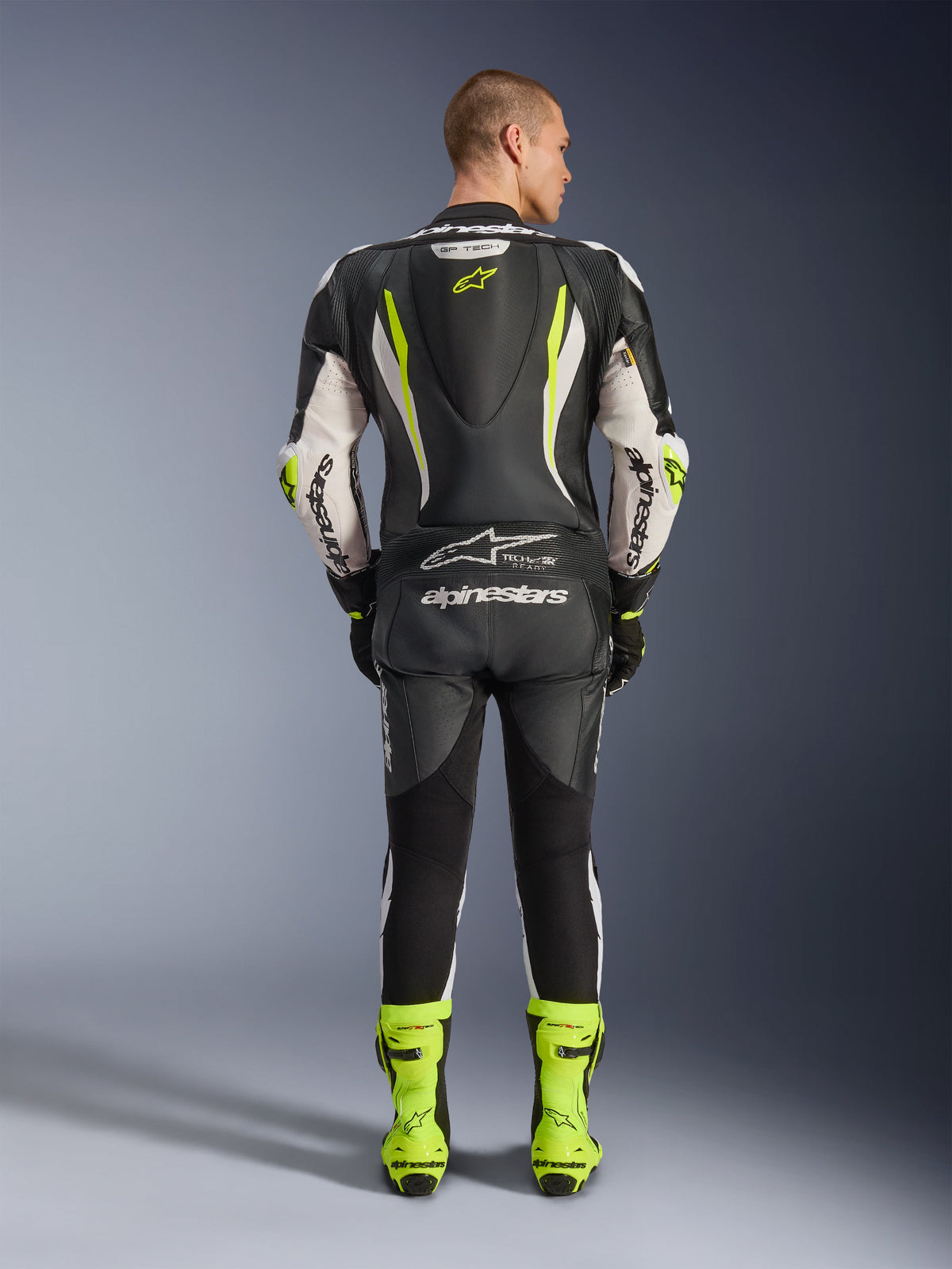 Alpinestars GP Tech V5 One Piece Leather Suit, Rear view, Black White and Yellow Fluo, featuring an aerodynamic speed hump with yellow star logo, white Alpinestars branding on sleeves and back, matching neon yellow racing boots, professional motorcycle track gear