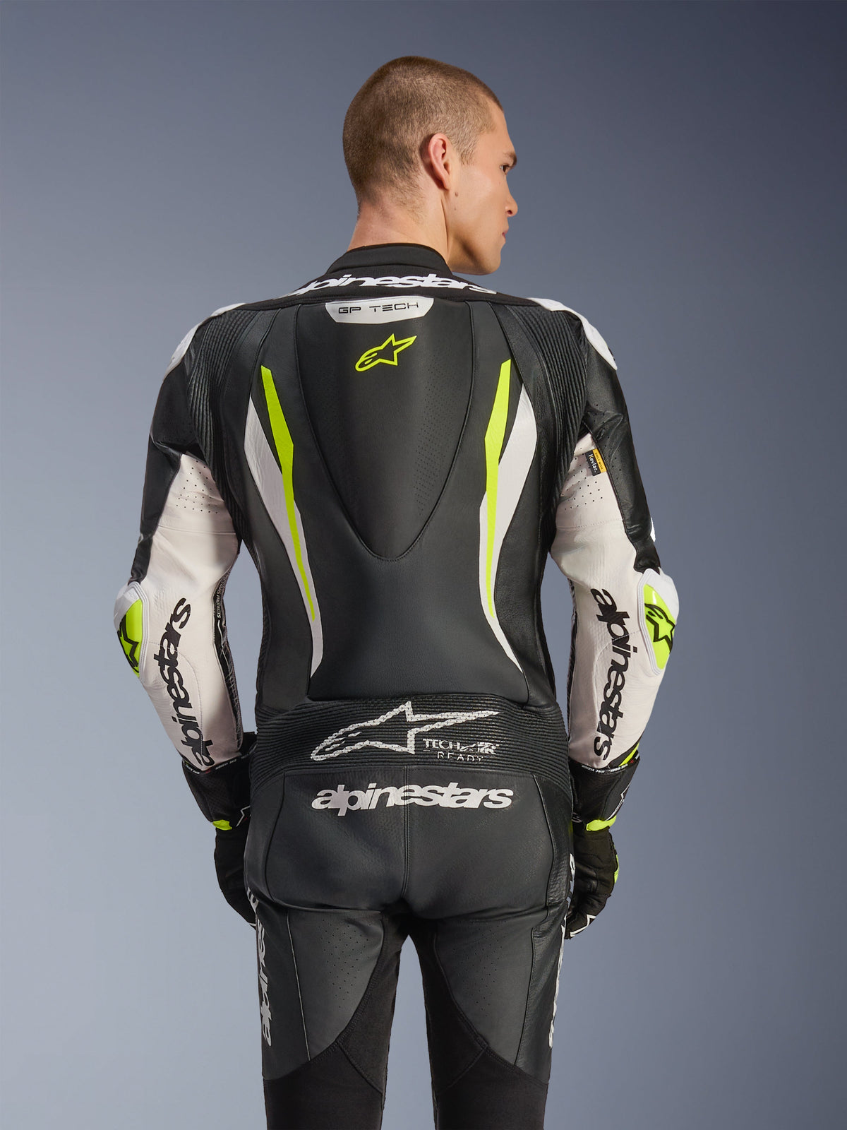 Alpinestars GP Tech V5 One Piece Leather Suit, back view on model, Black White and Yellow Fluo, featuring aerodynamic hump, large Alpinestars branding on arms and seat, accordion stretch panels, and Tech-Air Ready logo