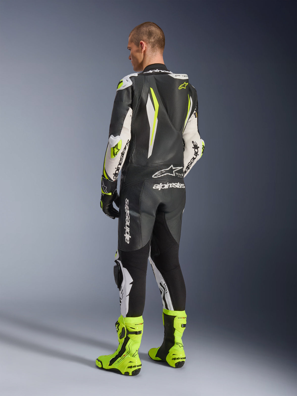 GP Tech V5 One Piece Leather Suit, Black White and Yellow Fluo, back view of rider wearing full racing suit with matching fluorescent yellow boots, featuring an aerodynamic speed hump and bold Alpinestars logos, professional motorcycle gear