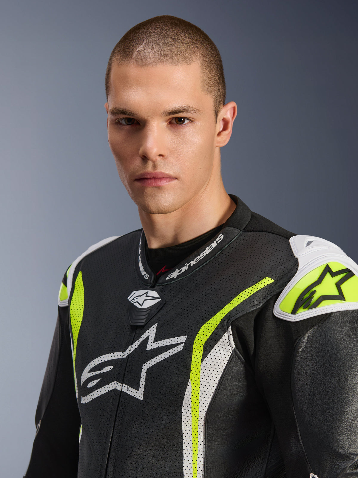 Close-up of a male model wearing the Alpinestars GP Tech V5 One Piece Leather Suit, Black White Yellow Fluo, featuring perforated leather, white Alpinestars logos on the chest and shoulders, and vibrant yellow accents, professional racing gear.