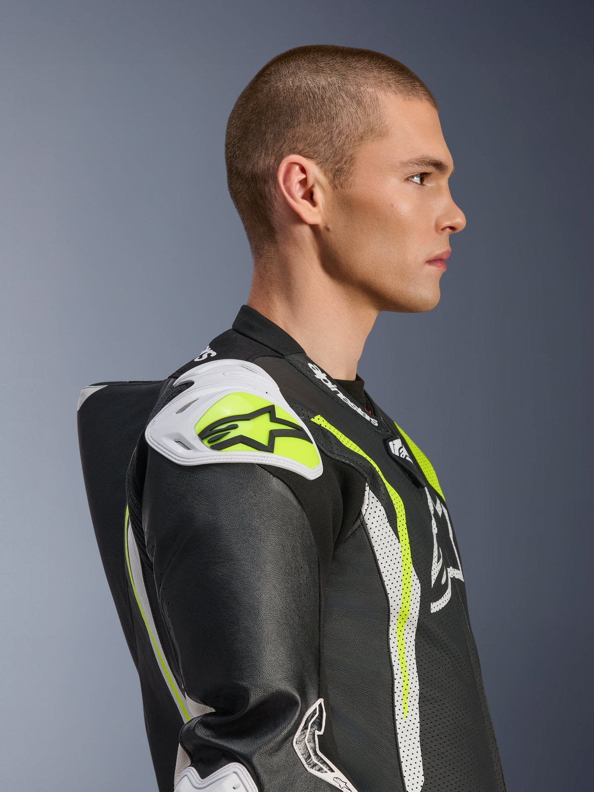 Male model wearing the Alpinestars GP Tech V5 One Piece Leather Suit in Black White Yellow Fluo, side profile view highlighting the aerodynamic speed hump and white shoulder sliders, racing-inspired motorcycle protective gear with perforated leather details