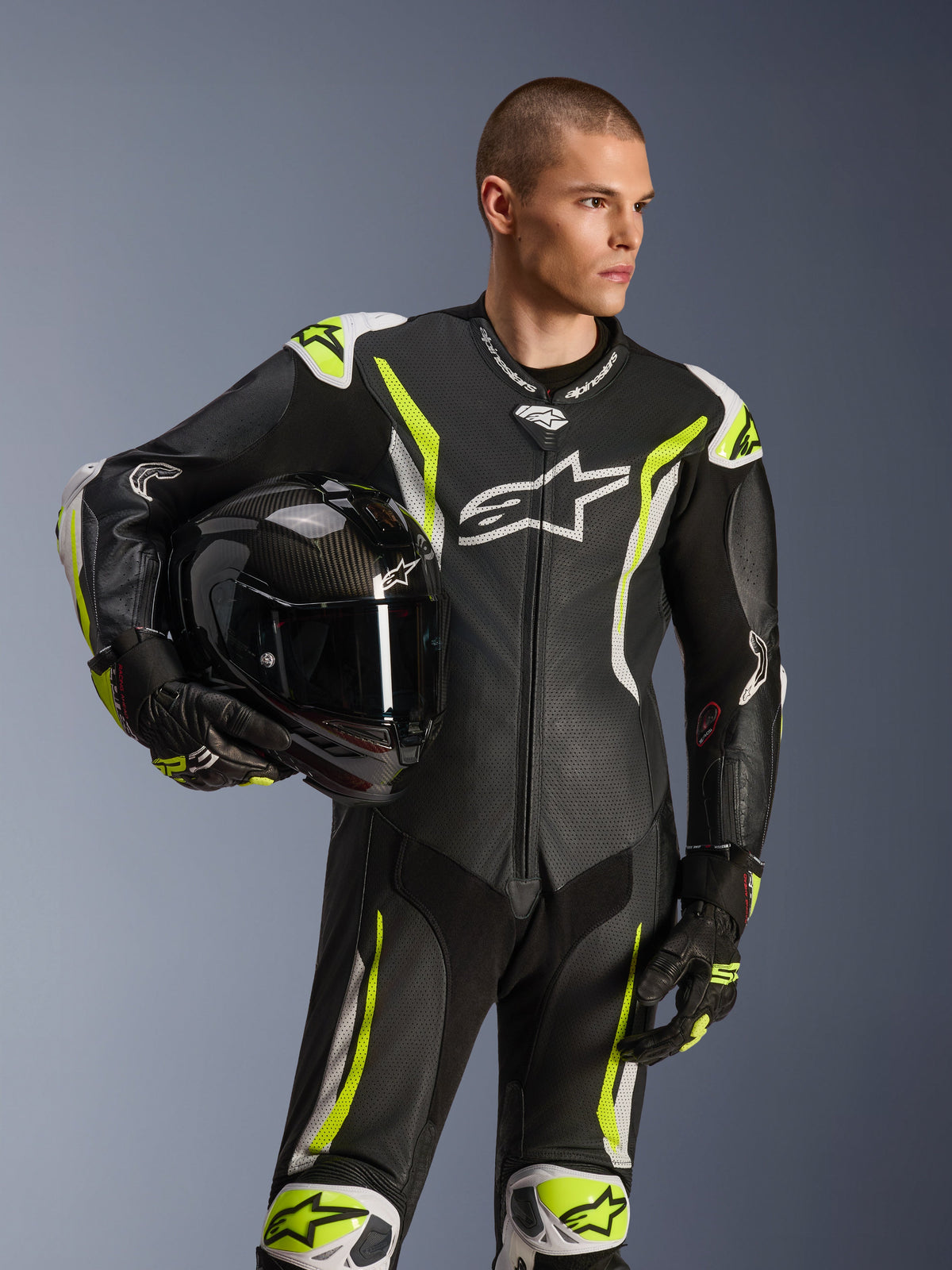 Alpinestars GP Tech V5 One Piece Leather Suit, Black White and Yellow Fluo, male model holding a black motorcycle helmet, featuring perforated panels, white and fluorescent yellow accents, and prominent Alpinestars branding, professional motorcycle racing gear