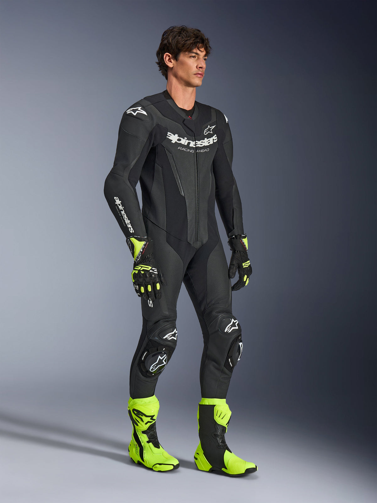 Man modeling the Alpinestars GP Force V2 Leather Suit 1-Piece in Black, featuring white Alpinestars branding, perforated leather sections, and integrated armor, styled with neon yellow and black racing boots and gloves