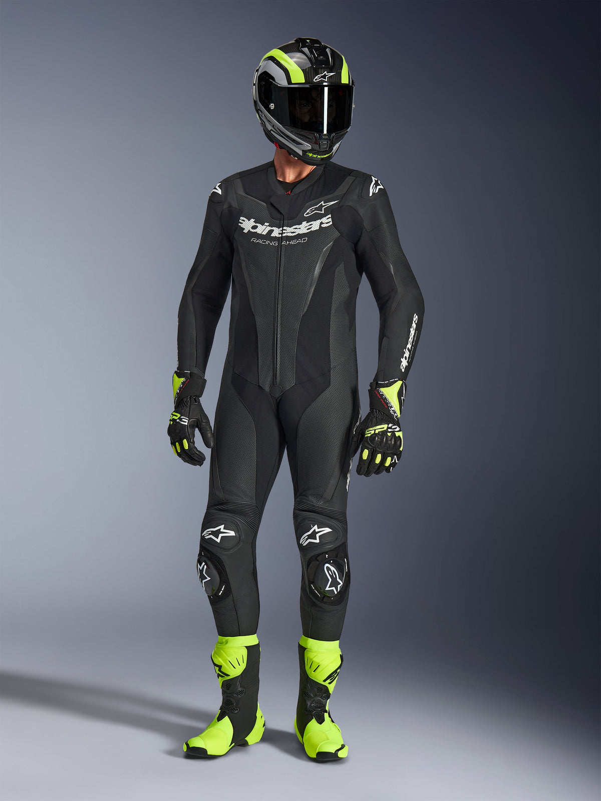 Motorcycle rider wearing the Alpinestars GP Force V2 Leather Suit in Black, 1-piece leather suit with white branding, paired with a black and fluo yellow racing helmet, gloves, and boots, professional track gear on a grey studio background