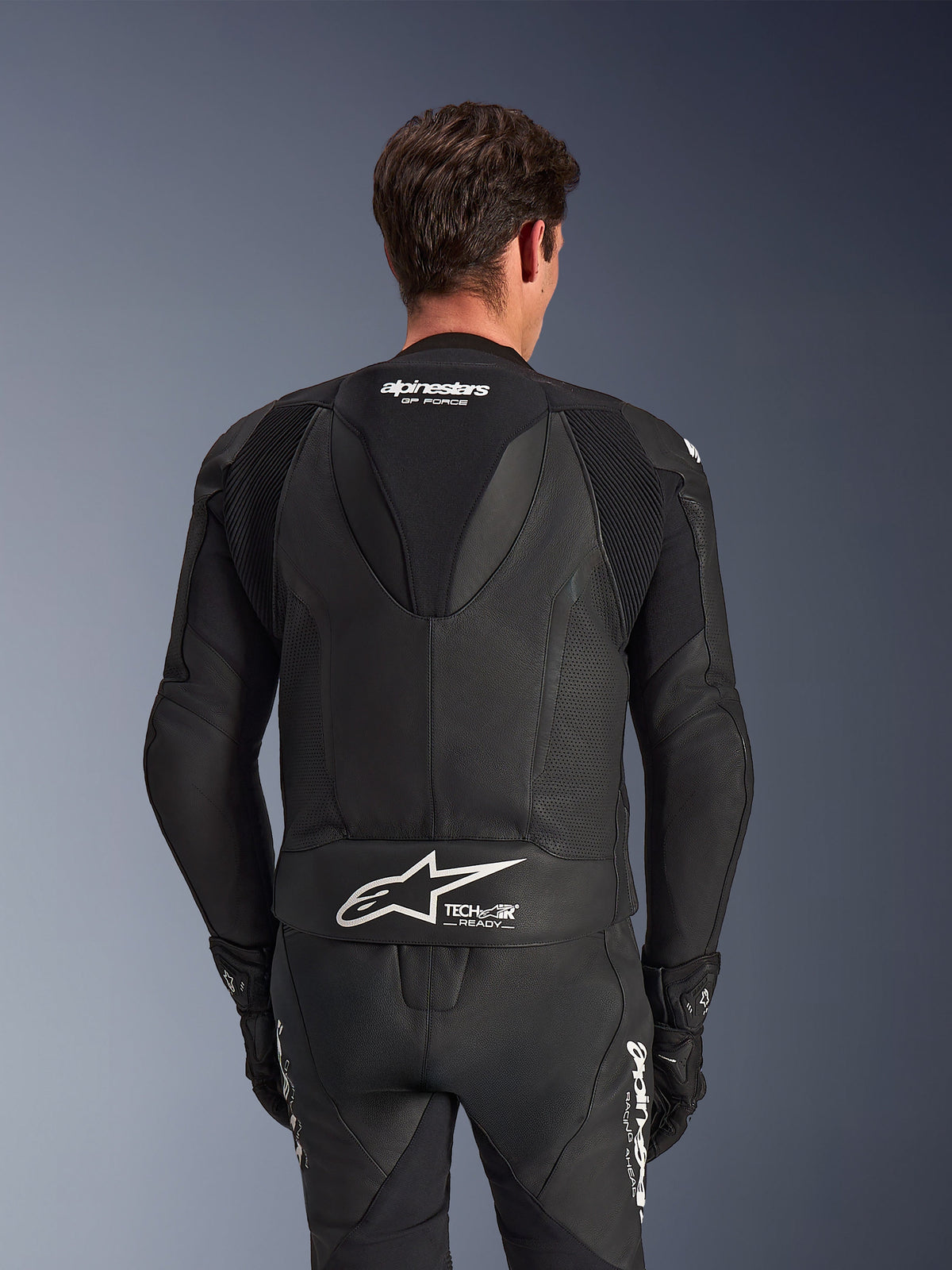Alpinestars GP Force V2 Leather Suit- 2Piece, Black, back view of rider, featuring perforated leather panels, aerodynamic speed hump with GP Force branding, and white Alpinestars logo with Tech-Air Ready text on the lower back.