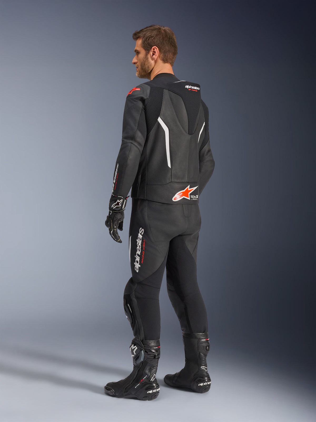 Alpinestars GP Force V2 2-Piece Leather Suit, Black with White and Red Fluo accents, rear three-quarter view on model, featuring an aerodynamic speed hump, perforated leather panels, and bold Alpinestars branding, worn with black racing boots and gloves.