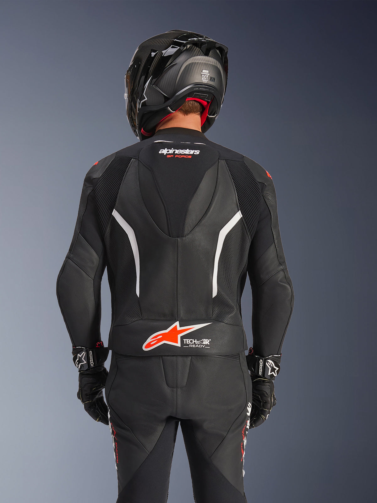 Rear view of Alpinestars GP Force V2 2-Piece Leather Suit in Black, featuring perforated leather, white accents, and red Alpinestars branding on the lower back, worn with a black motorcycle helmet and racing gloves.