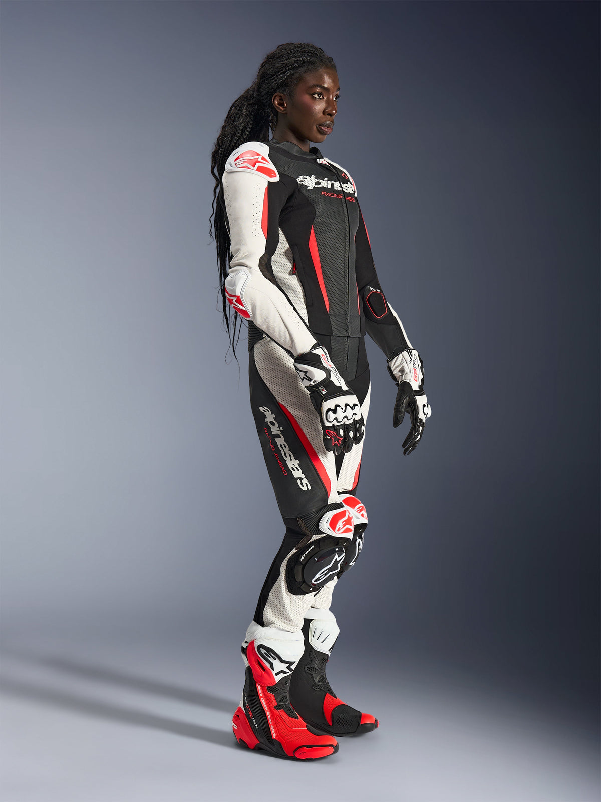 Kvinnlig förare iklädd Alpinestars Stella GP-R7 2-delat läderställ i svart, vitt och röd fluo, kombinerat med matchande racinghandskar och stövlar, med perforerade läderpaneler och skydd på axlar, armbågar och knän, stående i en studiomiljö.