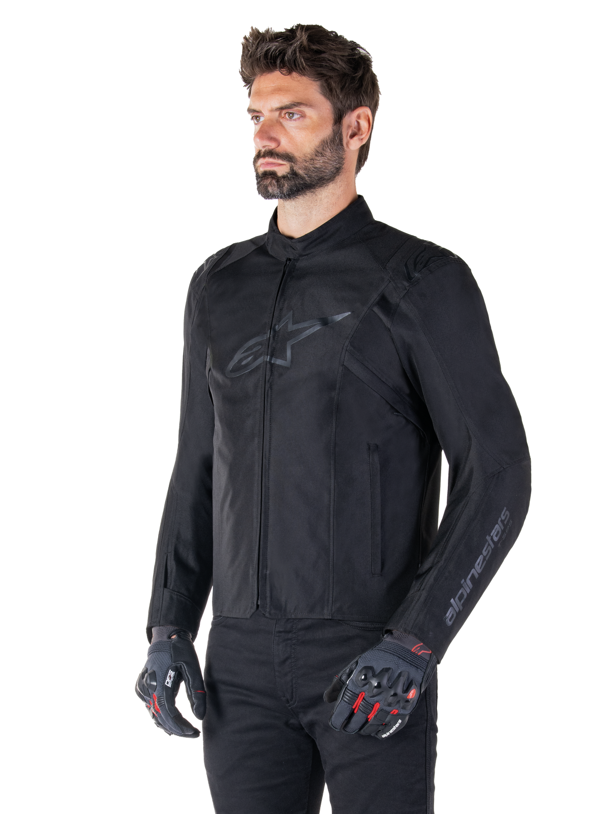 Modell som bär Alpinestars T-SPS V2 Waterproof Jacket, svart, kombinerad med svarta byxor och grå motorcykelhandskar med röda detaljer, med en diskret svart bröstlogotyp och logotyp på ärmen, sportig textiljacka för vägkörning.