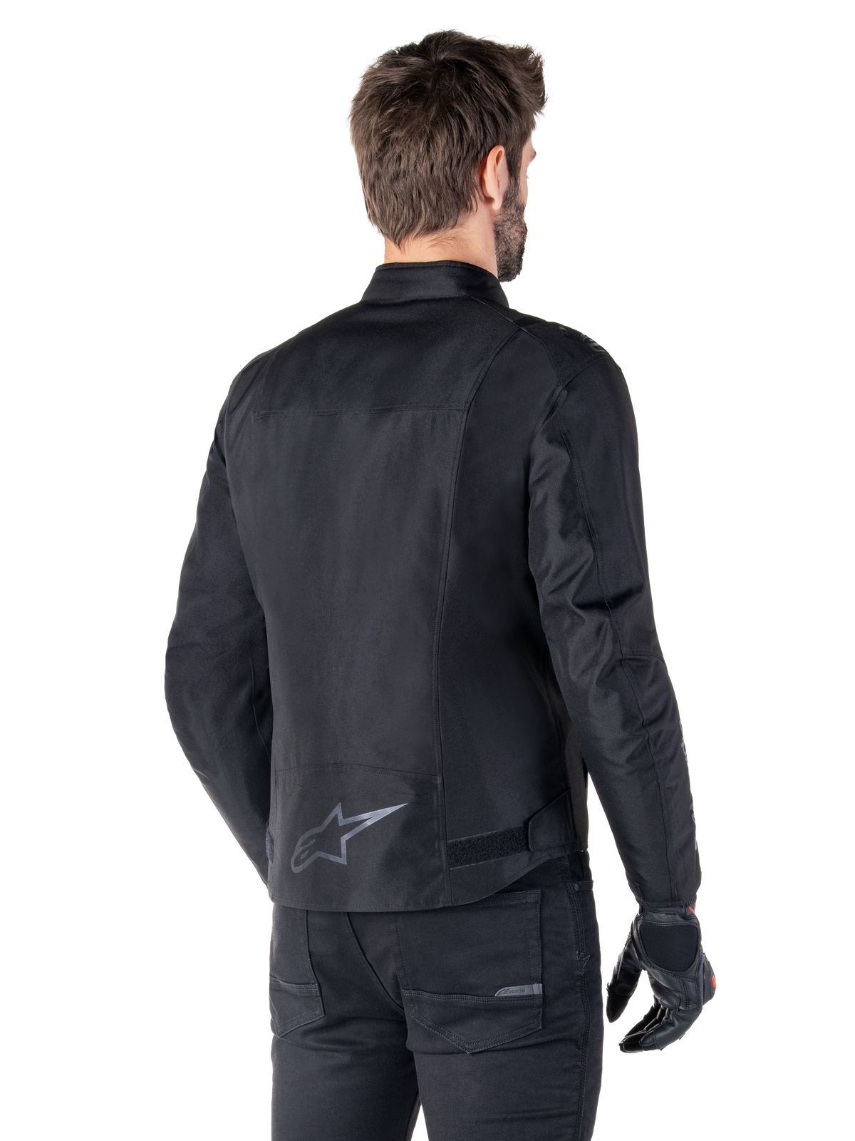 Alpinestars T-SPS V2 Vattentät Jacka, svart, sedd bakifrån på en manlig modell, med en reflekterande stjärnlogotyp på nedre delen av ryggen, förlängt ryggparti och justeringsremmar i midjan, kombinerad med svarta mc-byxor och handskar