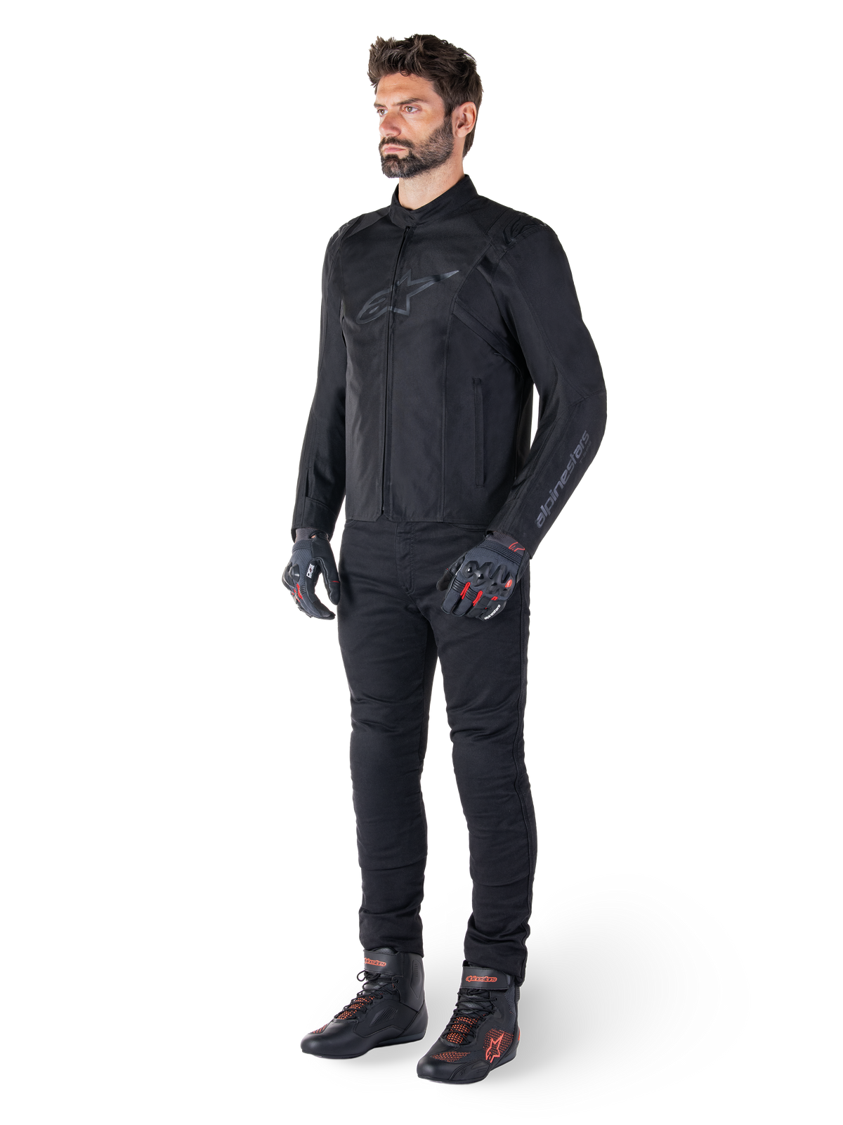 Alpinestars T-SPS V2 Vattentät Jacka, svart, bärs av en manlig modell med matchande svarta byxor, svarta handskar med röda detaljer och svarta mc-skor, komplett motorcykelutrustning för stads- och sportkörning, ren vit bakgrund