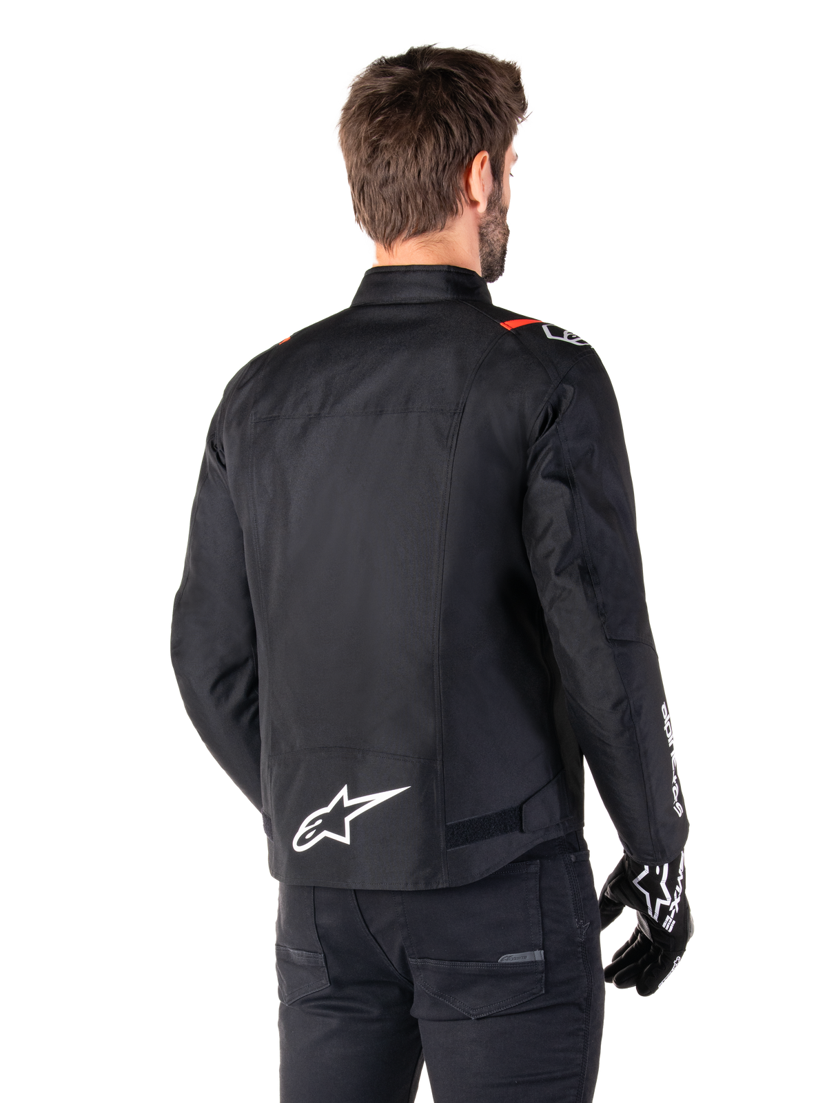 Alpinestars T-SPS V2 Waterproof-jacka, Black White Red Fluo, sedd bakifrån, med vit Alpinestars-logotyp på nedre delen av ryggen och röda detaljer på axlarna, bärs med svarta körhandskar och byxor, sportig textiljacka för motorcykel.