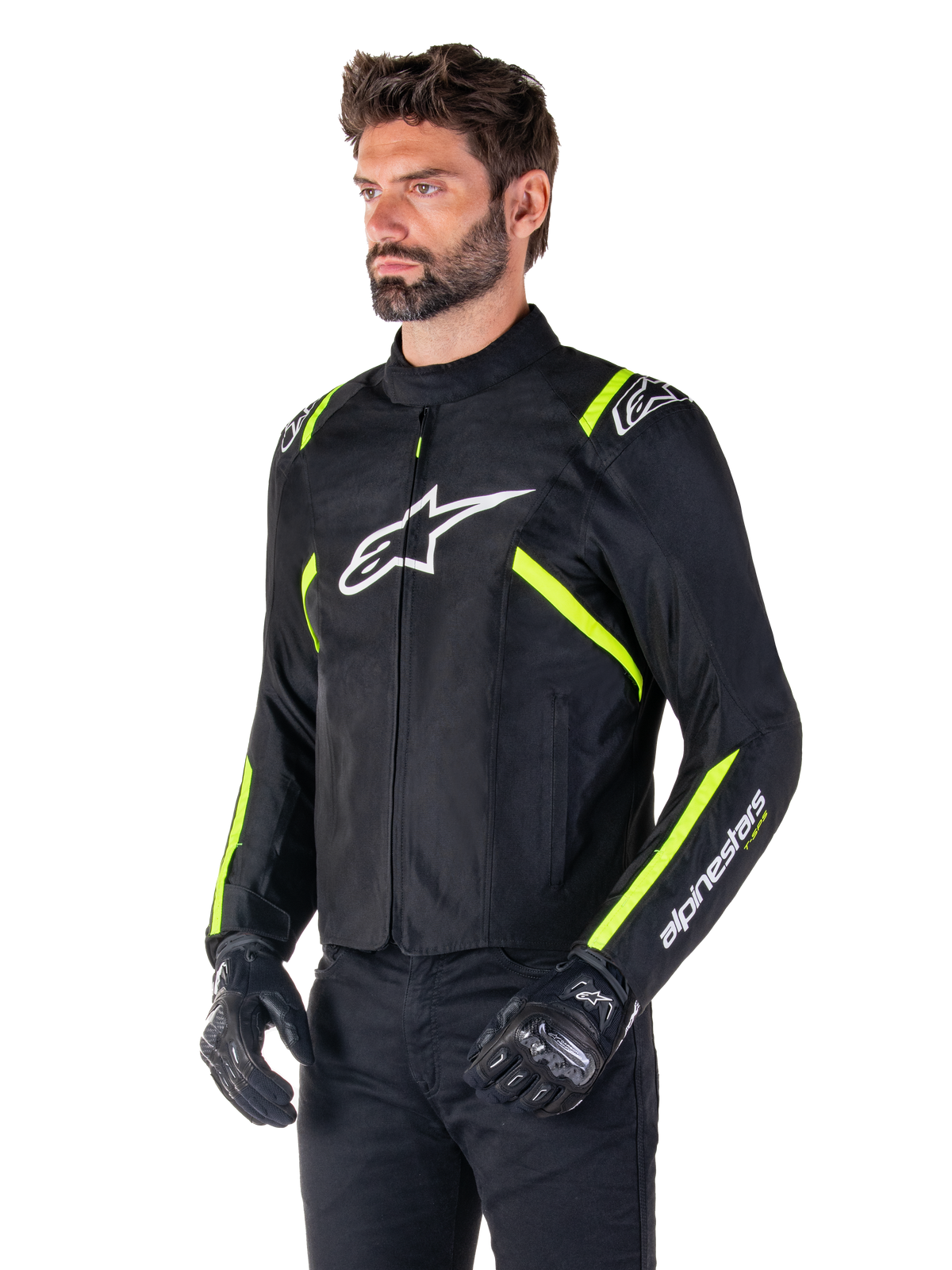 Manlig modell bär Alpinestars T-SPS V2 Waterproof Jacket, svart med vita och neongula detaljer, vit Astars-logotyp på bröstet och fluorescerande gula ränder på axlar och ärmar, svarta byxor och körhandskar, sportig textiljacka för motorcykel.