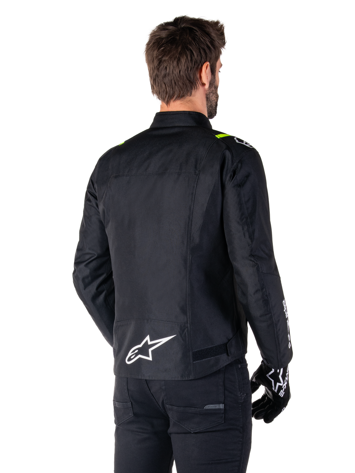Alpinestars T-SPS V2 Waterproof Jacket, svart vit gul fluo, bakifrån vy av manlig modell som bär motorcykeljackan i textil, med vit logotyp på nedre delen av ryggen och neongula detaljer på axlarna, kombinerat med svarta byxor och handskar.