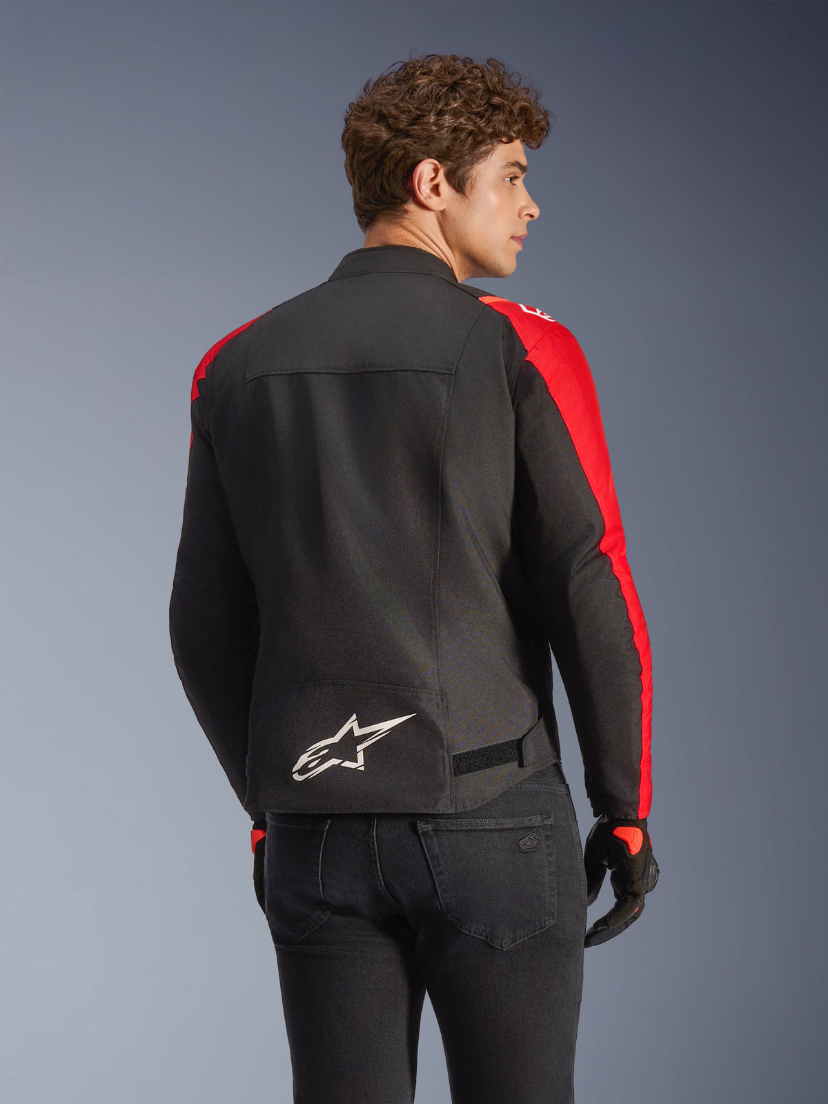 Alpinestars T-SPS V2 vattentät jacka i svart och rött, sedd bakifrån på en modell, med en vit Alpinestars-logotyp på nedre delen av ryggen, livfulla röda paneler på ärmarna och en sportig silhuett, bärs med svarta handskar och jeans.