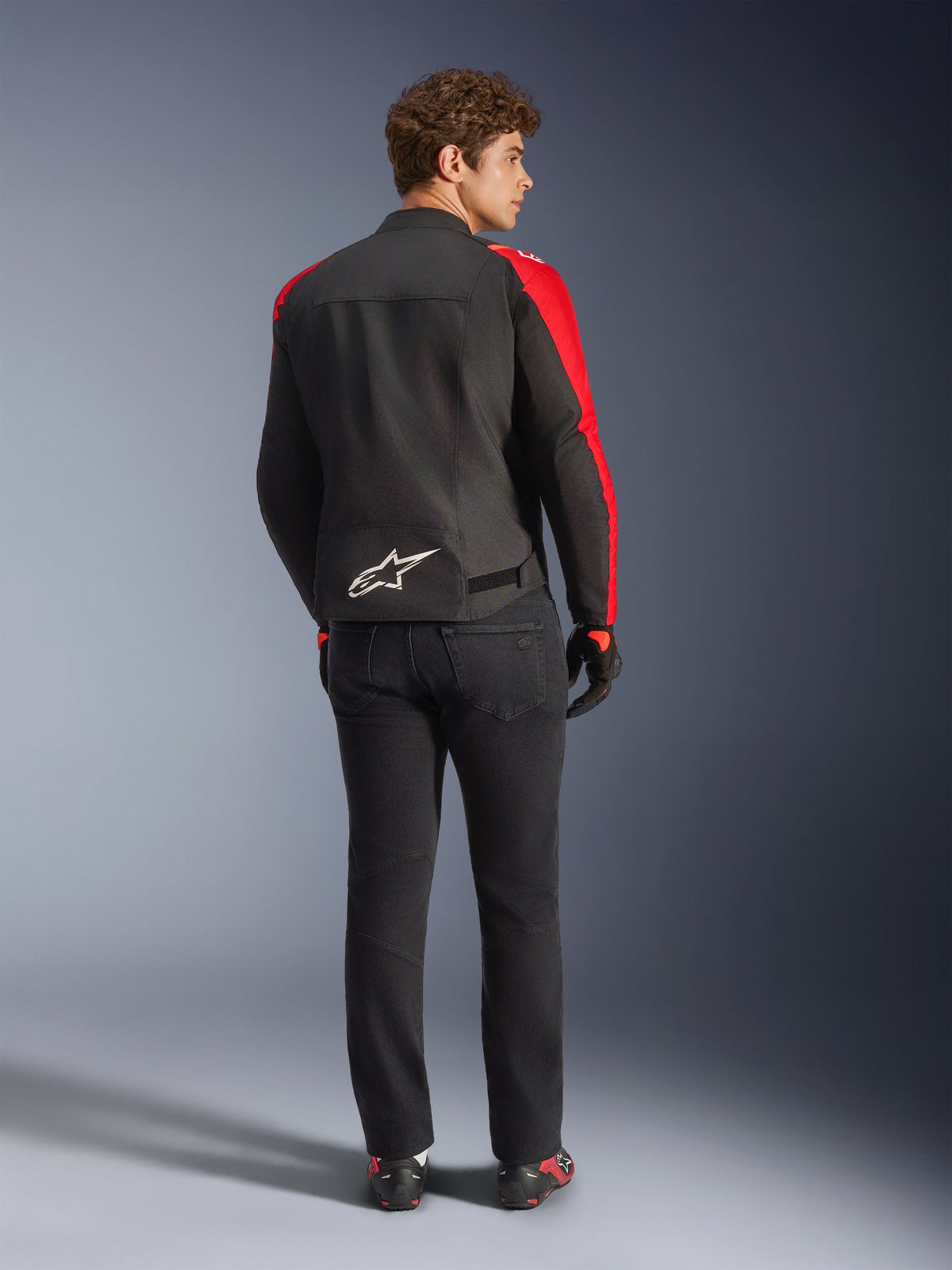 Alpinestars T-SPS V2 Waterproof Jacket, Black Red Red Fluo, vy bakifrån på manlig modell, vit Alpinestars-logotyp på nedre delen av ryggen, bärs med svarta motorcykelbyxor och handskar, sportig vattentät körjacka i textil