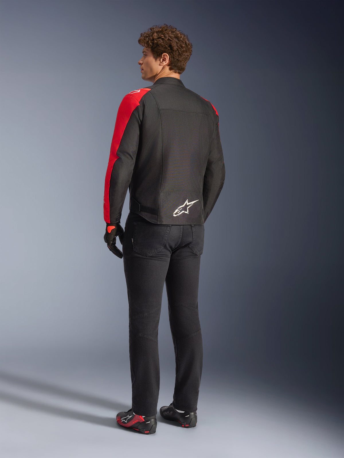 Alpinestars T-SPS V2 Waterproof-jacka i svart och röd fluo, sedd bakifrån på modell, bärs med svarta motorcykelbyxor och körskor, vit Alpinestars-logotyp på nedre delen av ryggen, textiljacka med sportig skärning
