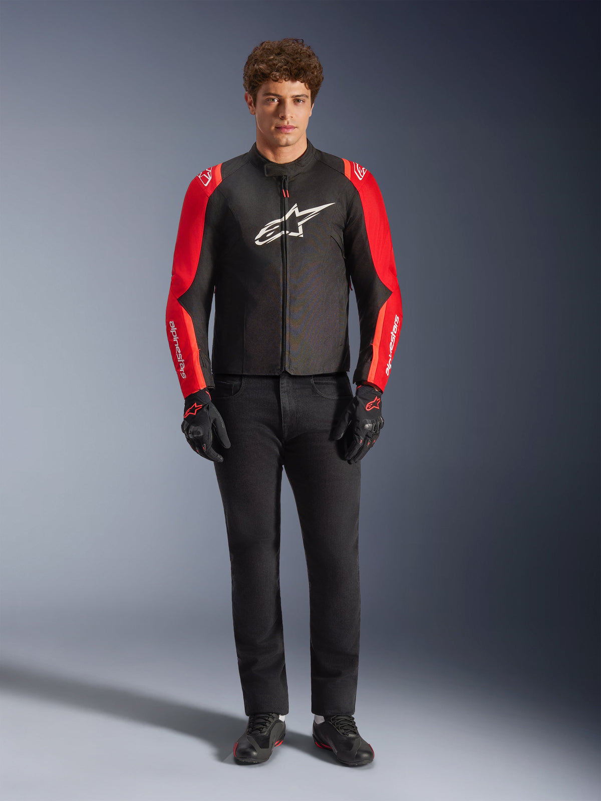 Manlig modell bär Alpinestars T-SPS V2 Waterproof-jacka, svart med röda och Red Fluo-ärmar, vit logotyp på bröstet, kombinerad med svarta byxor, svarta körhandskar och skor, sportig motorcykeldesign för vägkörning