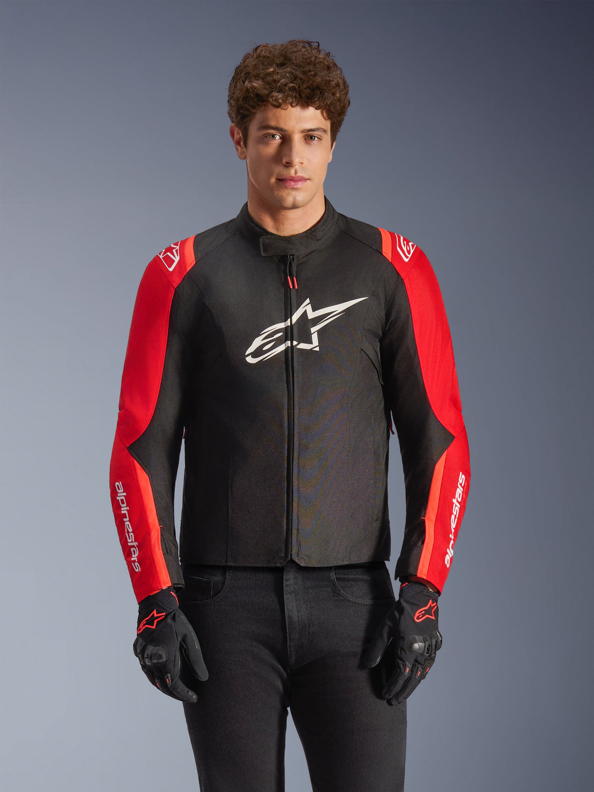 Alpinestars T-SPS V2 Waterproof Jacket, textil motorcykeljacka, svart och röd, bärs av en manlig modell med matchande svarta handskar, med en stor vit logotyp på bröstet och vita logotyper på röda ärmar, sportig silhuett för vägkörning i alla väder