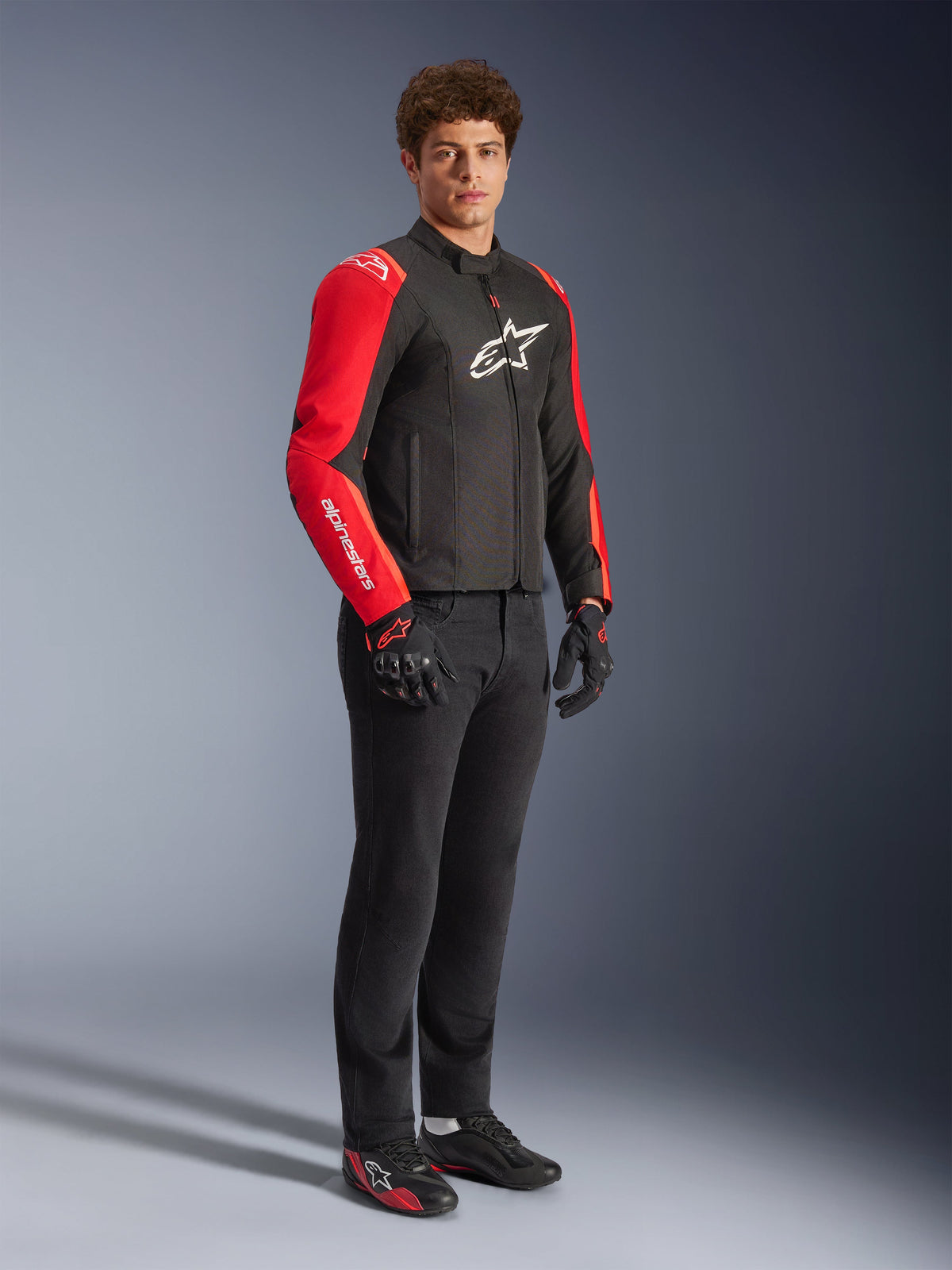 Manlig modell iklädd Alpinestars T-SPS V2 Waterproof-jacka i svart och Red Fluo, med klaröda ärmar med vita logotyper och en stor vit logotyp på bröstet, matchad med svarta mc-byxor, motorcykelhandskar och låga mc-skor, studiobild i helfigur.