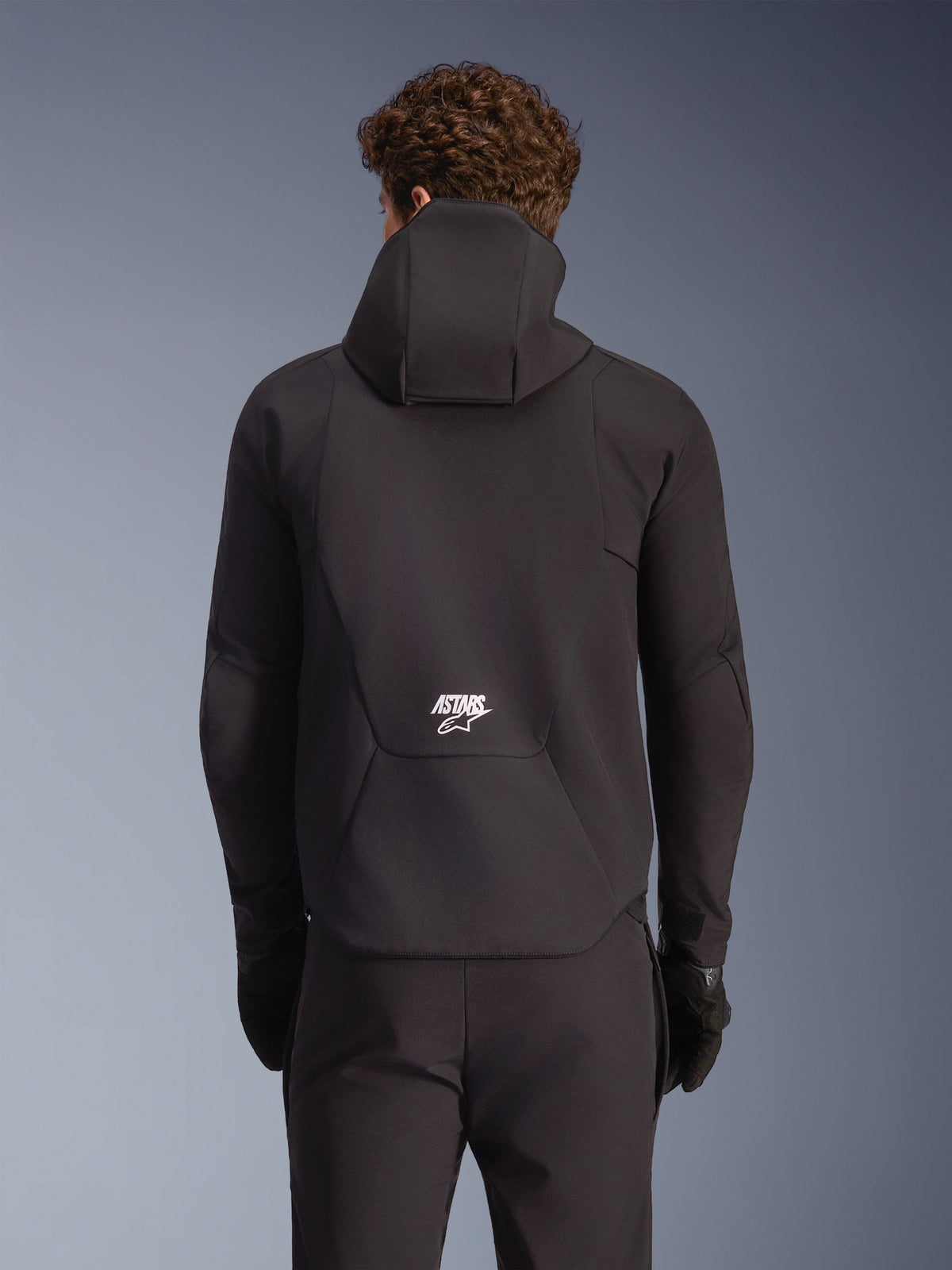 Alpinestars Aeroshell vattentät jacka, svart MC-softshelljacka, sedd bakifrån på modell med huva och vit ASTARS-logotyp.