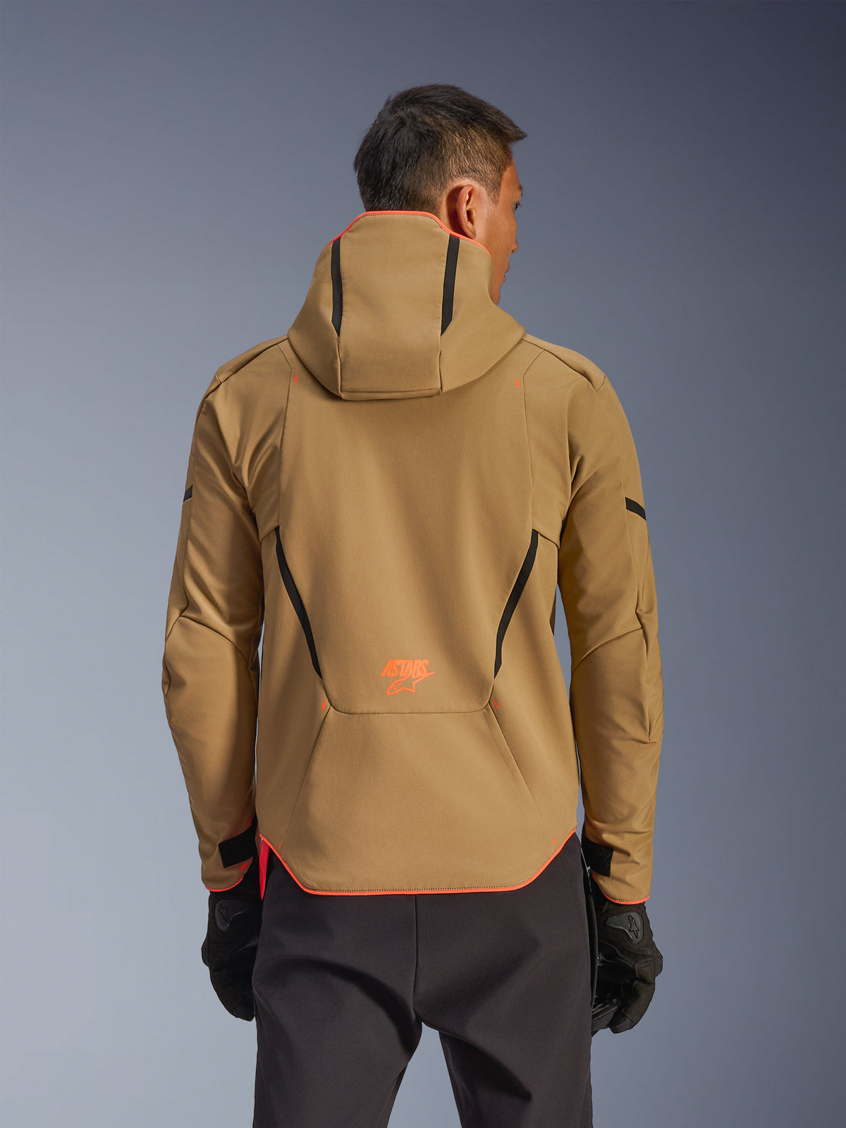 Alpinestars Aeroshell Waterproof Jacket, mörk khaki och orange fluo, baksida som visar huvtröjedesign med orange detaljer på huva och fåll, orange logotyp på nedre ryggen, bärs med svarta handskar, crossover sportig softshell-motorcykelutrustning