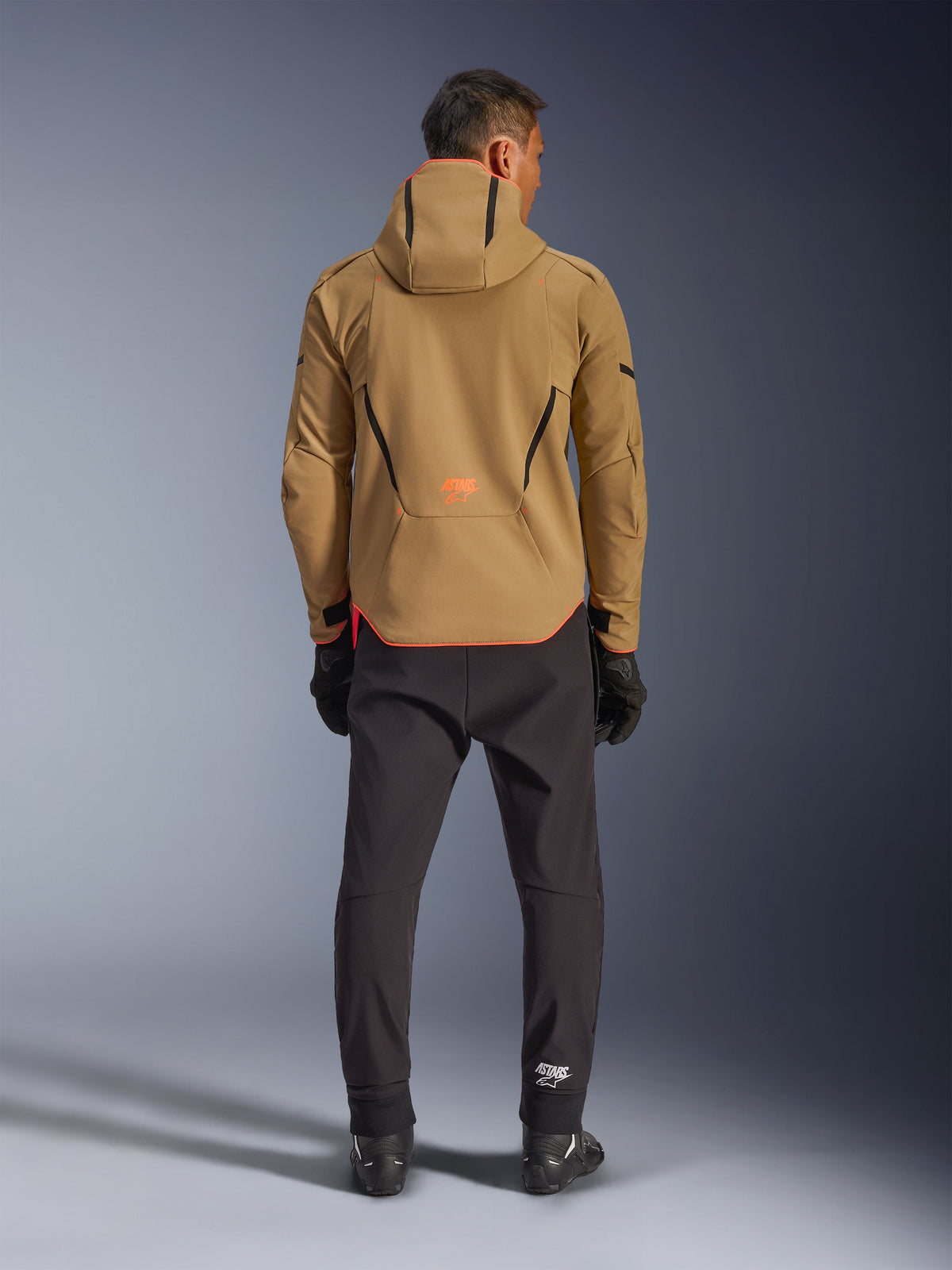 Alpinestars Aeroshell Waterproof Jacket i Dark Khaki Black Orange Fluo, baksida av en man som bär den huvförsedda softshelljackan, med svarta passpoaler och orange logotyp, kombinerad med svarta tekniska byxor och handskar.