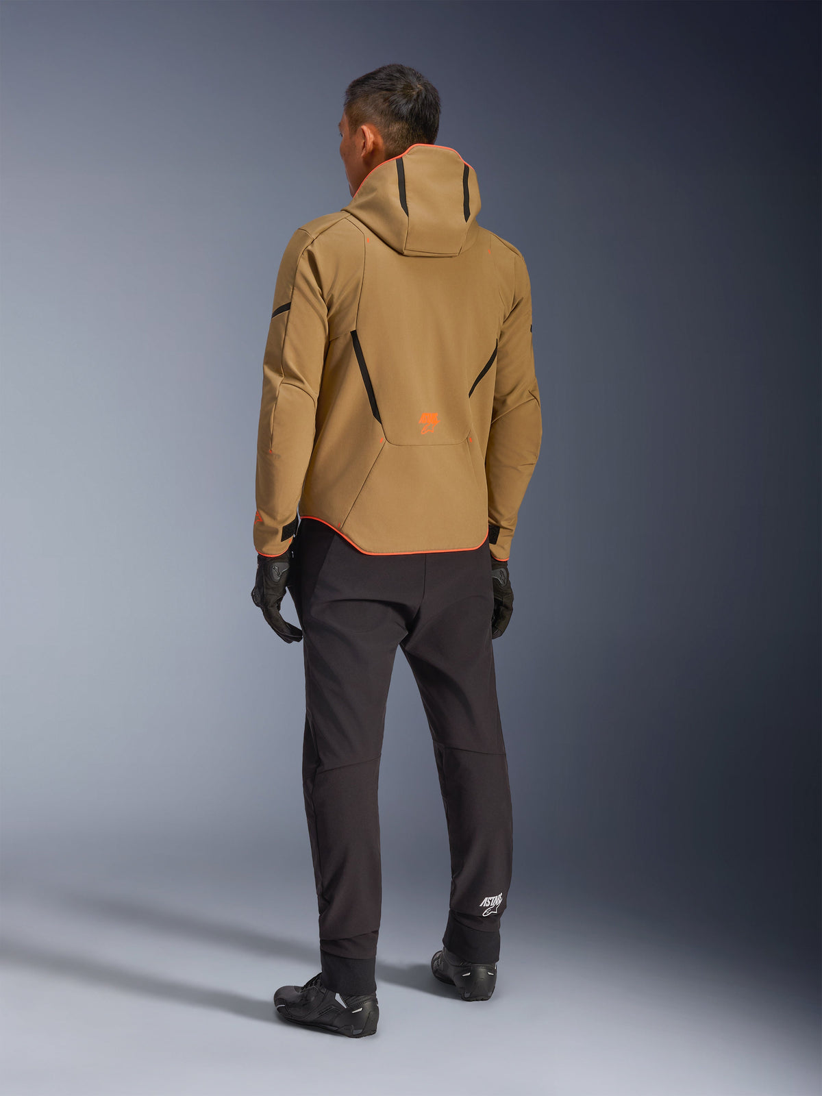 Alpinestars Aeroshell Waterproof Jacket, Dark Khaki Black Orange Fluo, vy bakifrån av en förare som bär den huvförsedda softshelljackan, matchad med svarta byxor och handskar, med svarta geometriska detaljer och en orange logotyp på nedre delen av ryggen.