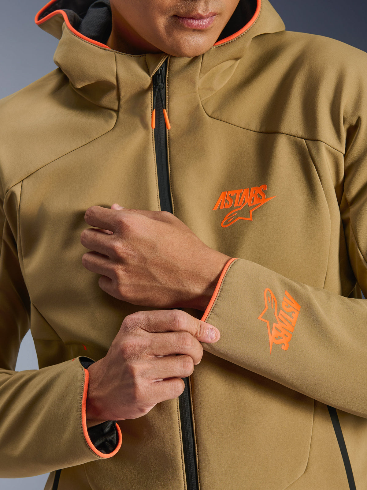 Alpinestars Aeroshell Waterproof Jacket, Dark Khaki och Orange Fluo, närbild av framsidan och ärmen med orangea ASTARS-logotyper, orangea kantband på huvan och ärmsluten samt en svart vattentät dragkedja.