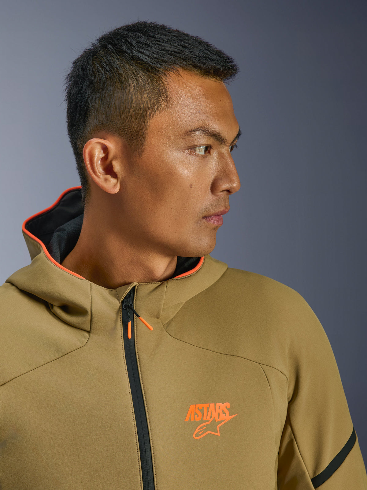 Alpinestars Aeroshell Waterproof Jacket, mörk khaki och orange fluo, närbild i sidoprofil, med orange ASTARS-logotyp på bröstet och orangefodrad huva, modern crossover-softshell-hoodie för motorcykelkörning