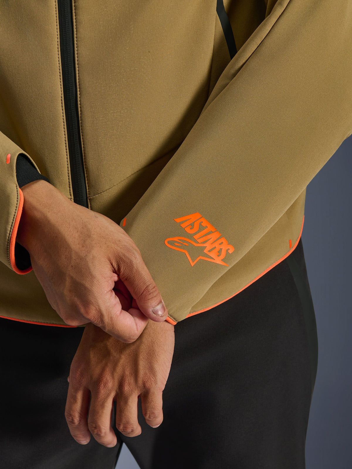 Alpinestars Aeroshell Waterproof Jacket, mörkt khakifärgad med neonorange detaljer, närbild på ärmslut och nederkant med den klarorangea Astars-logotypen, neonorangea kantband och vattentät dragkedja framtill, sportig softshell-konstruktion