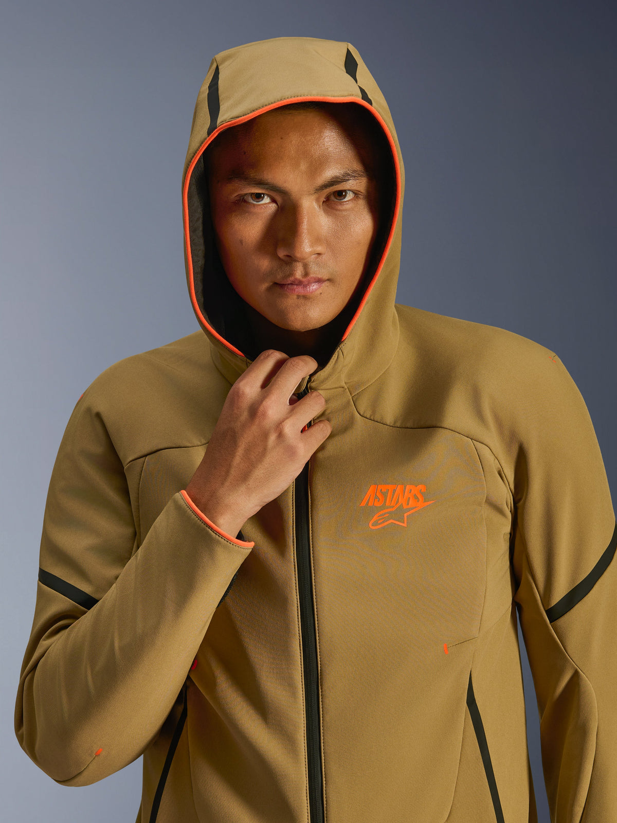 Alpinestars Aeroshell Waterproof Jacket, Dark Khaki Black Orange Fluo, modell som bär jackan med huvan uppe, neonorange detaljer och orange ASTARS-logotyp, svarta detaljer, modern sportig vattentät softshelljacka