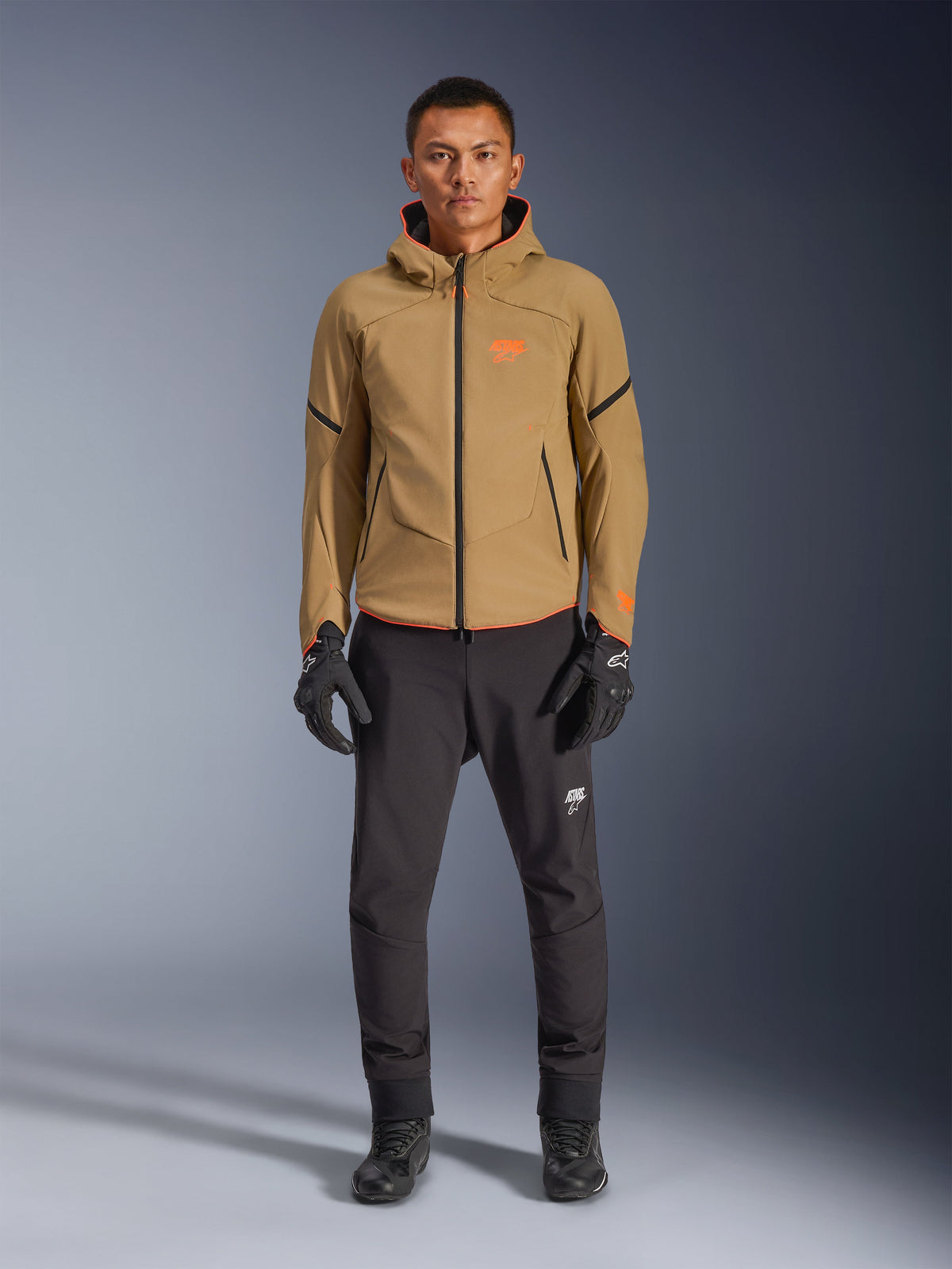 Manlig modell bär Alpinestars Aeroshell Waterproof Jacket i Dark Khaki med svarta och Orange Fluo-detaljer, orange logotyp på bröstet och design med huva, kombinerat med svarta byxor och svarta motorcykelhandskar, casual sport-touring-stil