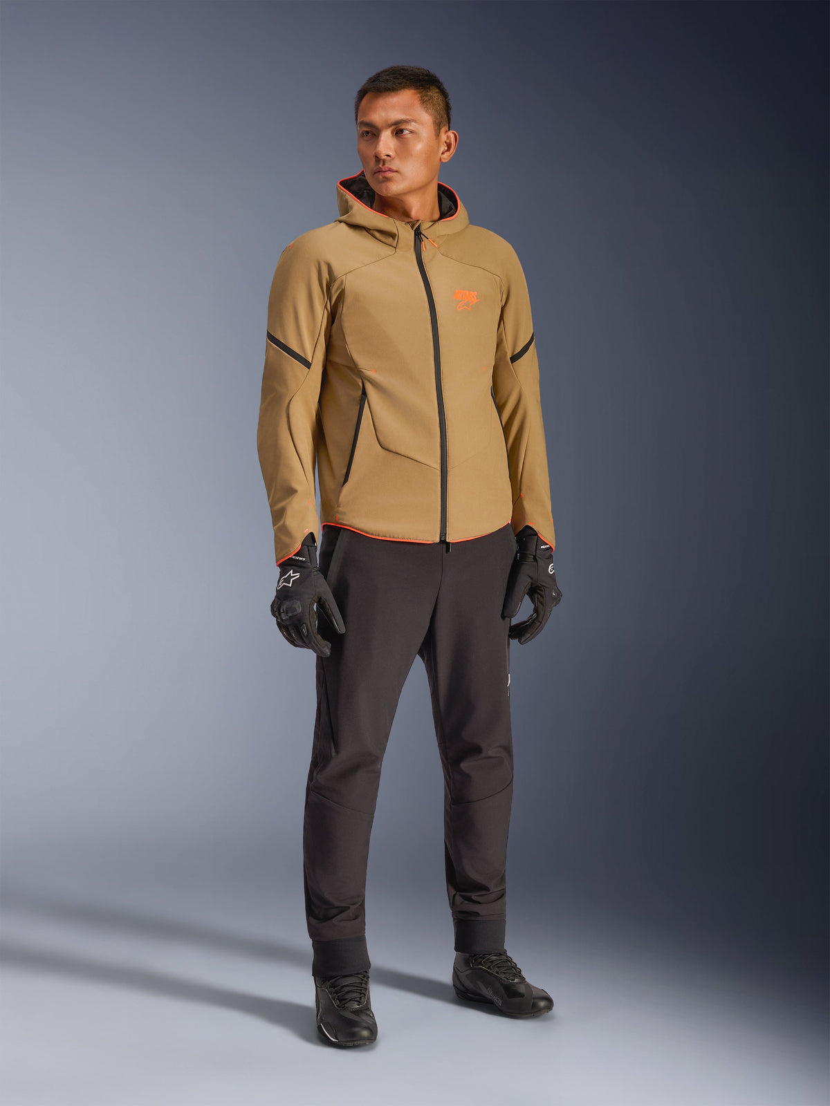 Alpinestars Aeroshell Waterproof Jacket i mörk khaki, svart och neonorange, bärs av en modell med matchande utrustning, sportig softshell-design med huva, vattentäta dragkedjor och detaljer i orange fluo