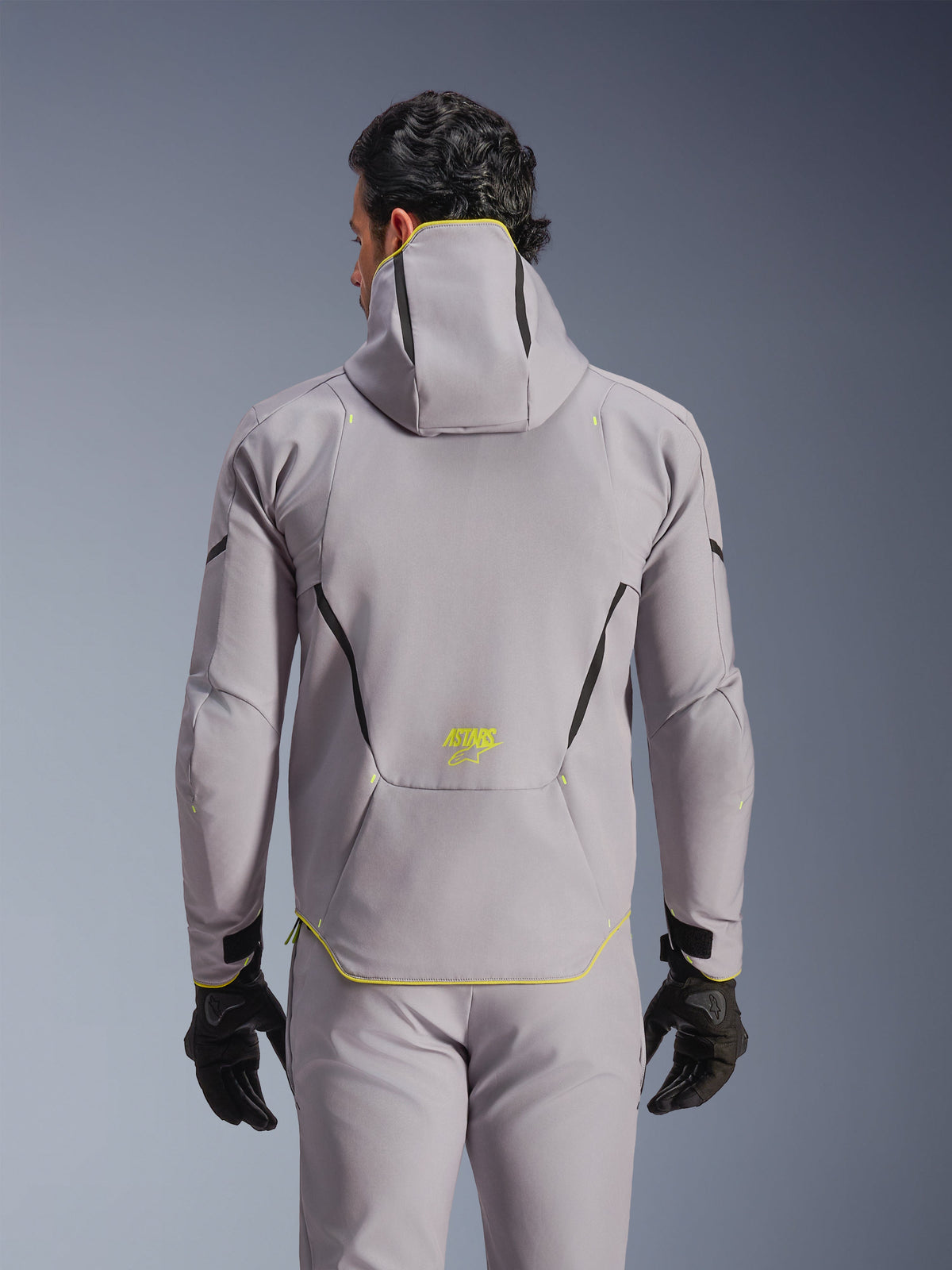 Alpinestars Aeroshell Waterproof Jacket, mellangrå med neongula detaljer, vy bakifrån som visar integrerad huva och svarta kontrastlinjer, gul ASTARS-logotyp på nedre delen av ryggen, bärs med matchande grå byxor och svarta handskar, sportig softshell-motorcykeljacka