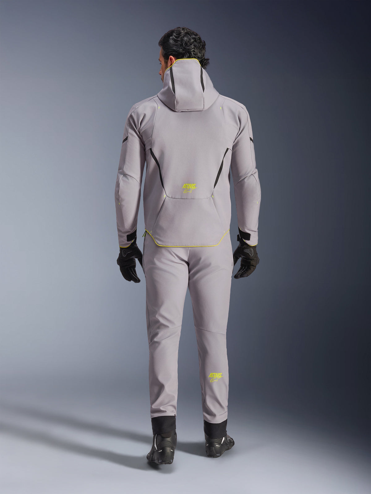 Alpinestars Aeroshell Waterproof Jacket i Mid Gray Yellow Fluo, vy bakifrån, bärs med matchande grå byxor, svarta handskar och svarta mc-skor. Har en softshelldesign med huva, fluorgula 'ASTARS'-detaljer och logotyper, konstruerad för modern motorcykelprestanda på väg.