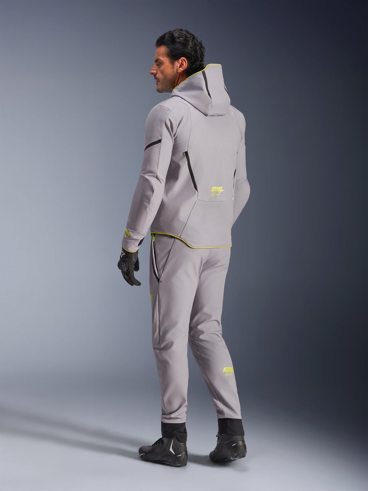 Alpinestars Aeroshell Waterproof Jacket, Mid Gray Yellow Fluo, bärs med matchande grå byxor, hoodie-design med gula detaljer och logotyper, vy bakifrån, kombinerad med svarta körhandskar och stövlar, modern motorcykelutrustning