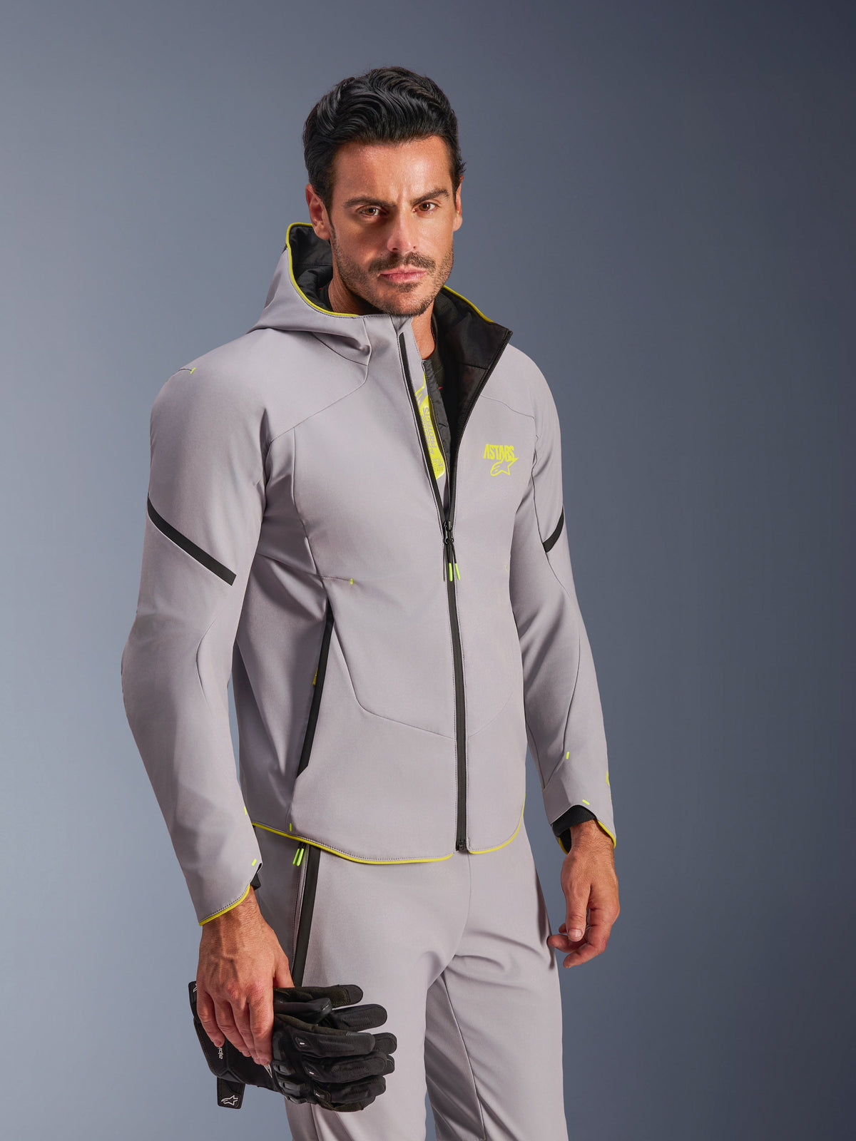 Alpinestars Aeroshell Waterproof Jacket i Mid Gray och Yellow Fluo, buren av en man med matchande grå byxor och svarta motorcykelhandskar, med en softshelldesign med huva, neongula detaljer och logotyp på bröstet, modern sportig motorcykelutrustning.