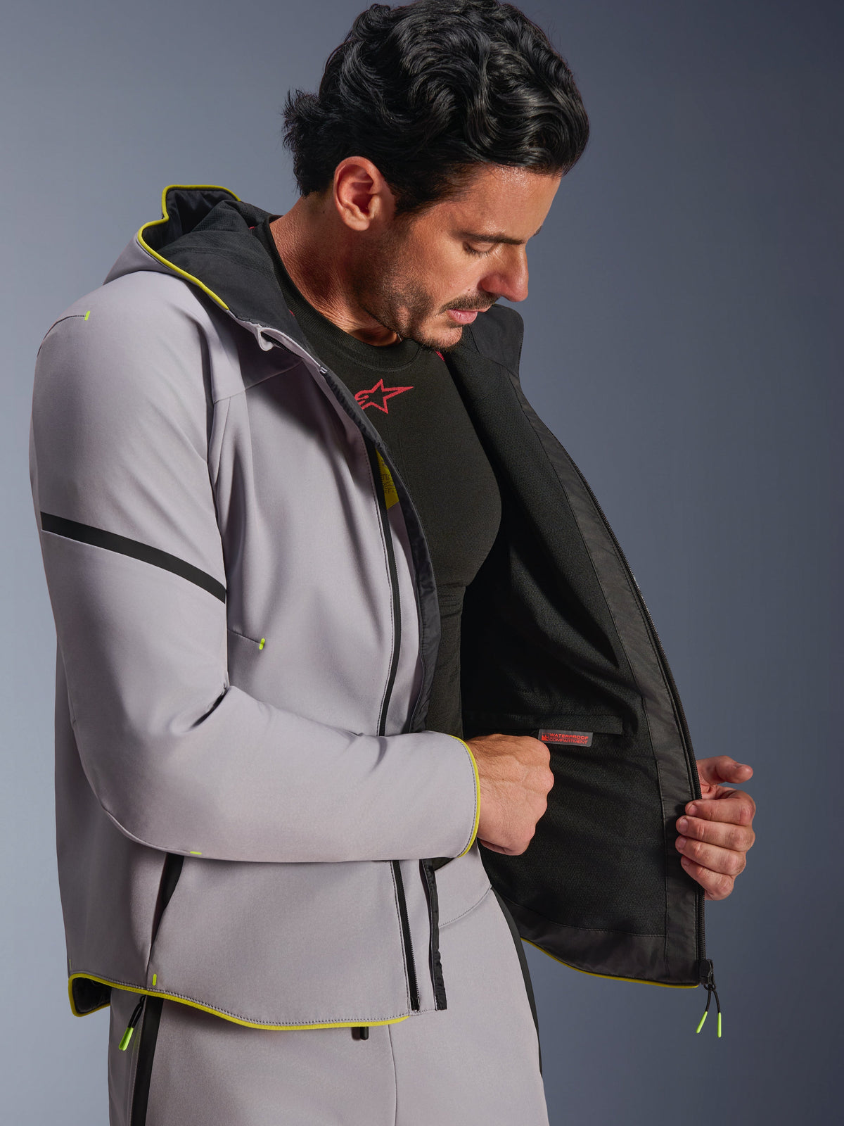 Alpinestars Aeroshell Waterproof Jacket i mellangrått och fluorescerande gult, buren av en manlig modell som visar det interna vattentäta facket och det svarta fodret, sportig softshell-motorcykelhoodie med fluorescerande gula detaljer och modern atletisk passform.