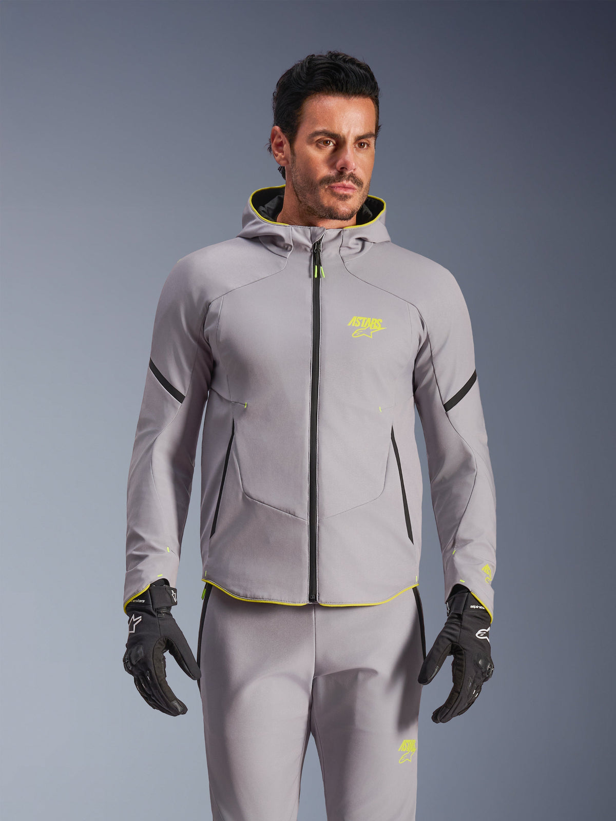 Alpinestars Aeroshell Waterproof Jacket, mellangrå och neongul, motorcykeljacka i softshell med huva och svarta dragkedjor, bärs med matchande grå byxor och svarta handskar, vattentät prestandautrustning för landsvägskörning