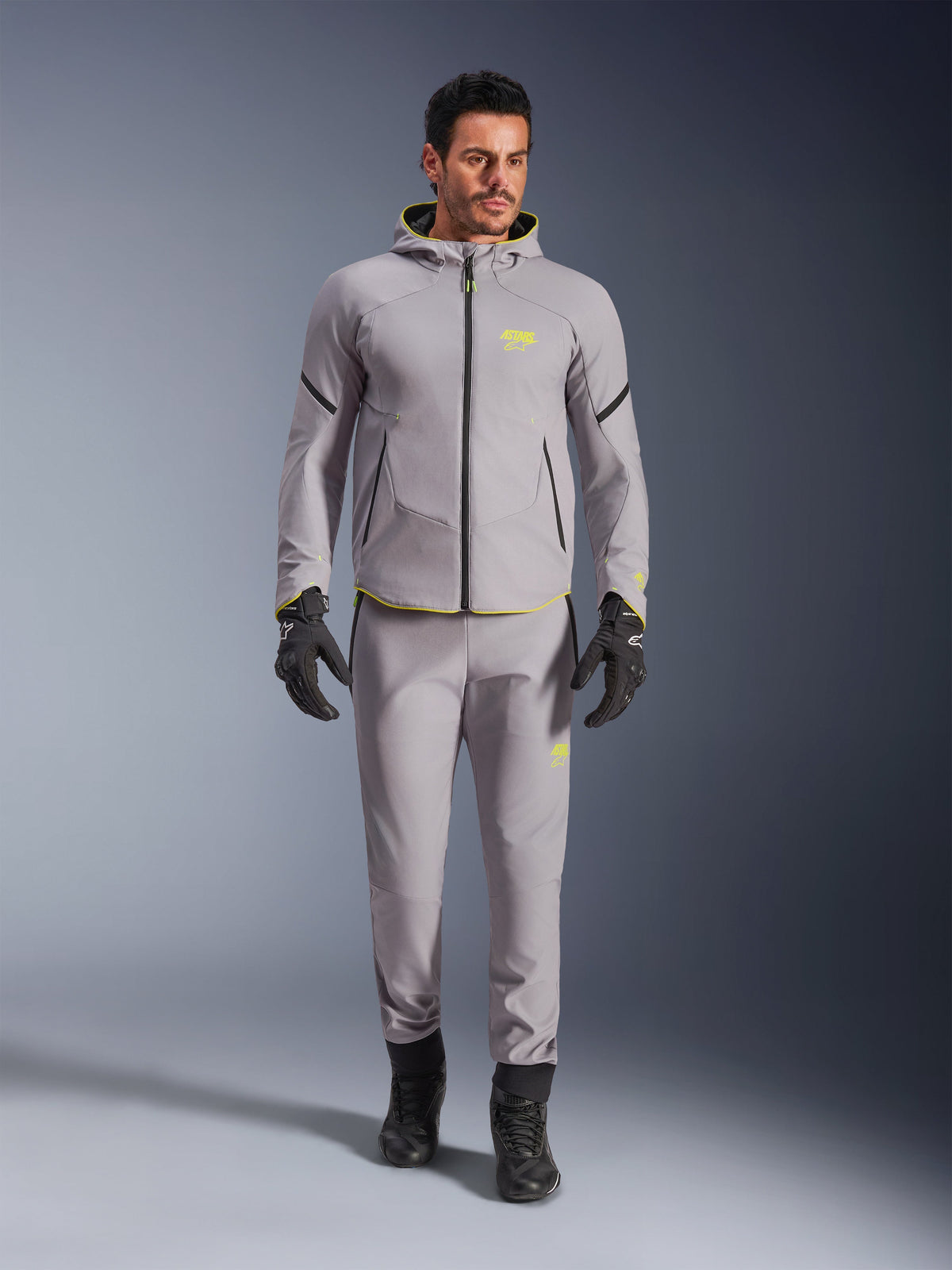 Alpinestars Aeroshell Waterproof Jacket, Mid Gray Yellow Fluo, buren med matchande grå byxor, svarta handskar och stövlar, med en modern sportinspirerad hoodie-design med livfulla gula detaljer och atletisk passform.