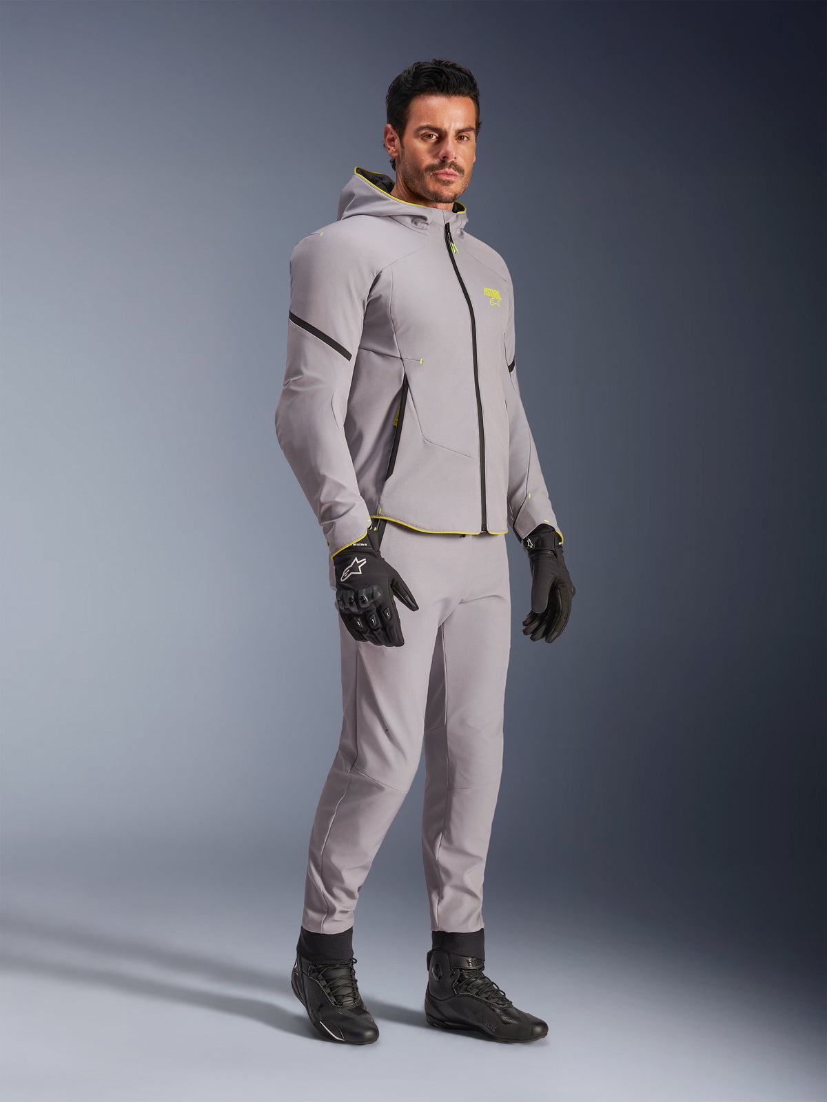 Alpinestars Aeroshell Waterproof Jacket, Mid Gray och Yellow Fluo, bärs av modell med matchande grå byxor, svarta körhandskar och svarta stövlar, modern motorcykelutrustning i hoodie-stil för vardaglig och sportig körning.