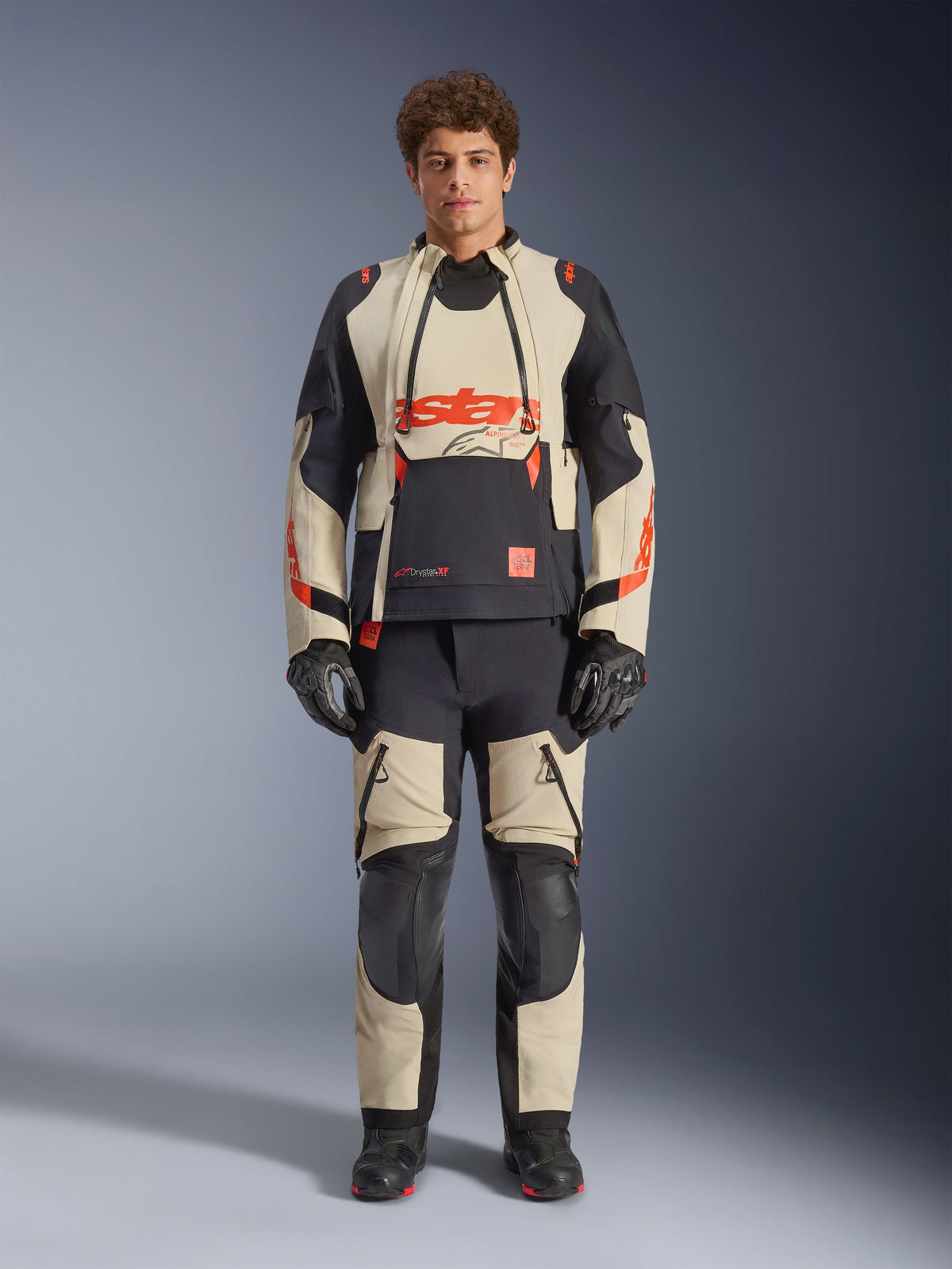 Alpinestars Halo Pro Drystar® XF laminerad jacka, Mastic Black, adventure-motorcykeljacka bärs av en modell med matchande byxor och handskar, beige och svarta textilpaneler med tydliga röda logotyper, modulär design för terrängkörning
