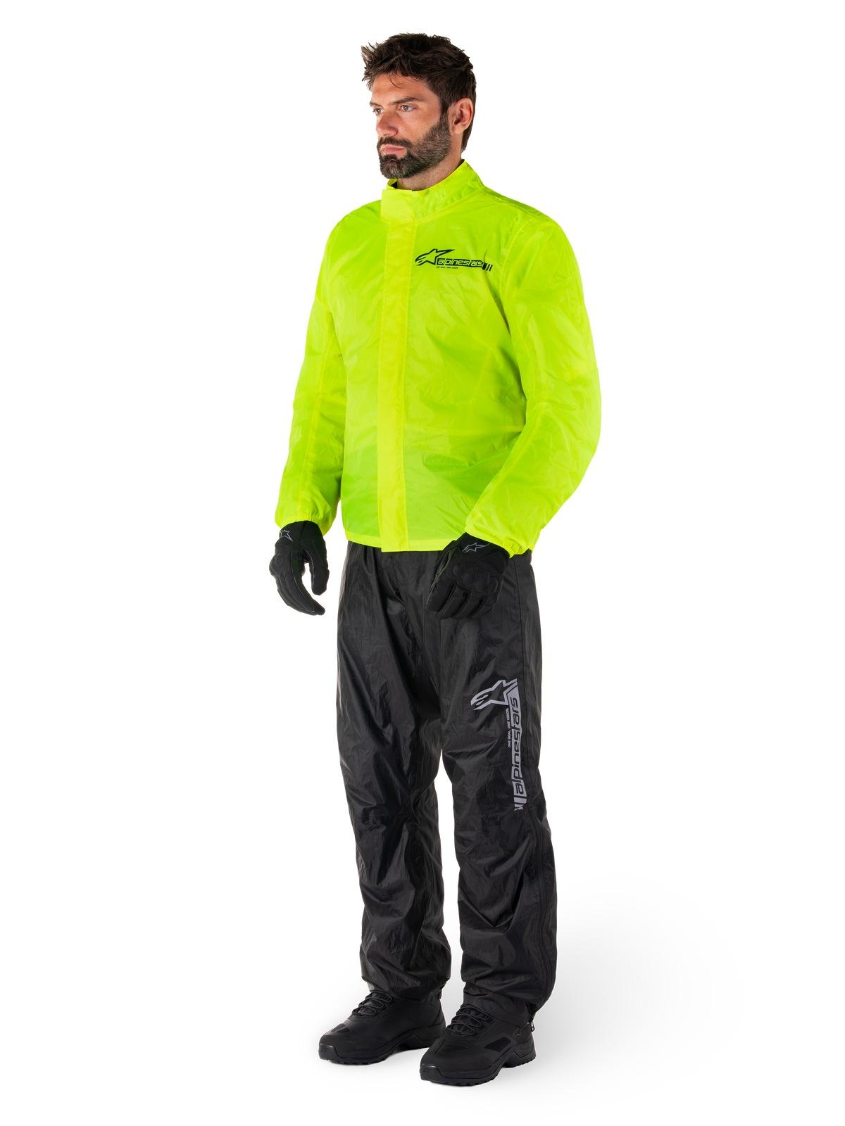 Man i Alpinestars Hurricane Rain V2-jacka i Yellow Fluo och Hurricane Rain V2-byxor i svart, vattentät motorcykelregnutrustning med hög synlighet, med reflekterande logotyper på bröst och lår, komplett outfit med svarta handskar och stövlar.