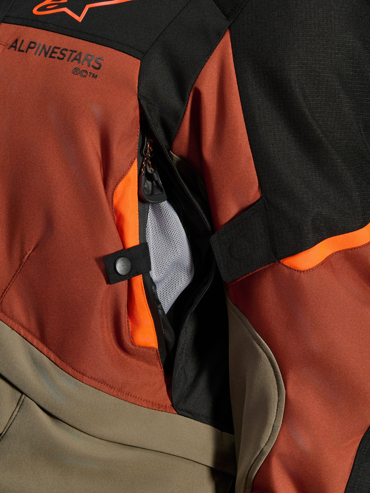 Alpinestars Andes V4 Drystar® Jacka, Walnut Rust Black, närbild på direkt ventilationsöppning med nätfoder, hållbart textilmaterial med orangea detaljer, tryckknapp och Alpinestars logotypdetalj