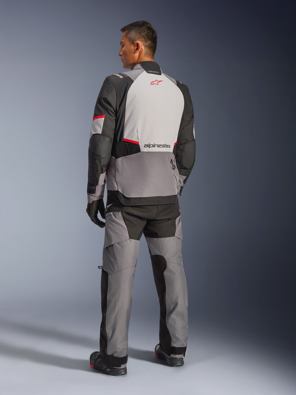 Alpinestars Andes V4 Drystar®-jacka, mörkgrå och ljusgrå med svarta och klaröda detaljer, vy bakifrån som visar en ljusgrå panel med röd stjärnlogotyp, kombinerad med matchande adventure-byxor och svarta handskar, teknisk motorcykelutrustning för alla väder.