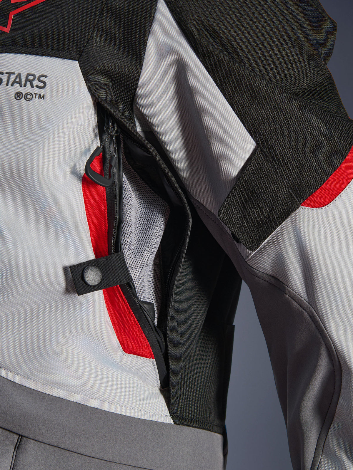 Närbild på detalj av Alpinestars Andes V4 Drystar®-jacka i mörkgrått, isgrått och klarrött, som framhäver en bröstventilationsöppning med dragkedja, nätfoder och en justeringsrem med tryckknapp.