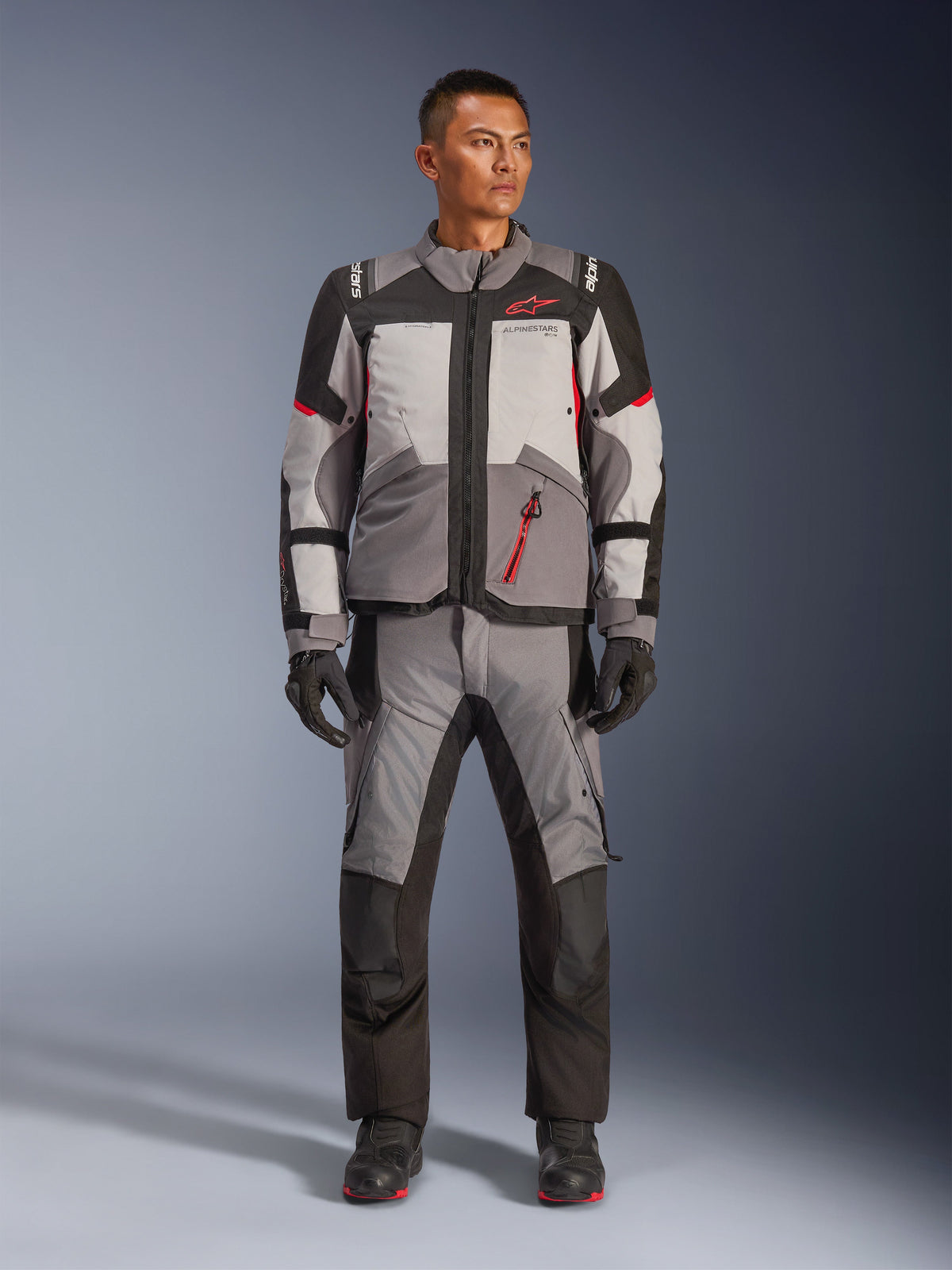 Alpinestars Andes V4 Drystar® Jacket, adventure-motorcykeljacka, mörkgrå, isgrå, svart och klarröd, bärs med matchande textilbyxor och handskar, med en teknisk flerpanelsdesign och röd Alpinestars-logotyp, adventure-touring-utrustning.